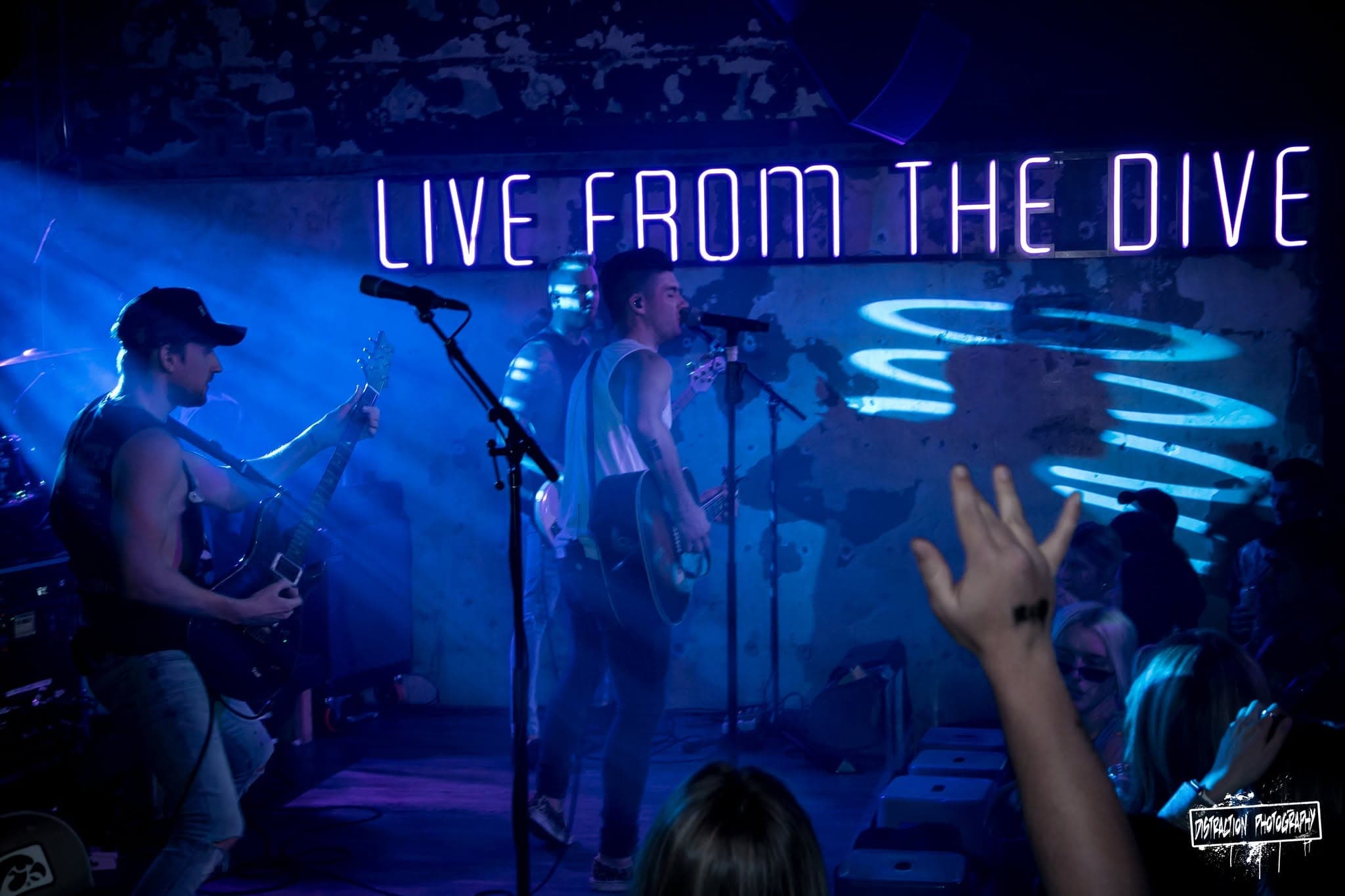 A band performing on stage under the neon 'Live From The Dive' sign at Elray's, with a crowd raising their hands up front