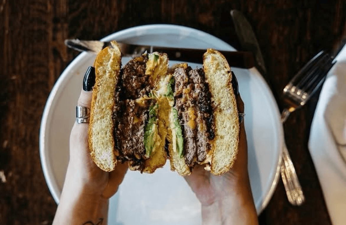 A hand holding up a thick, juicy smashed double cheeseburger with avocado, sliced in half to show the layers
