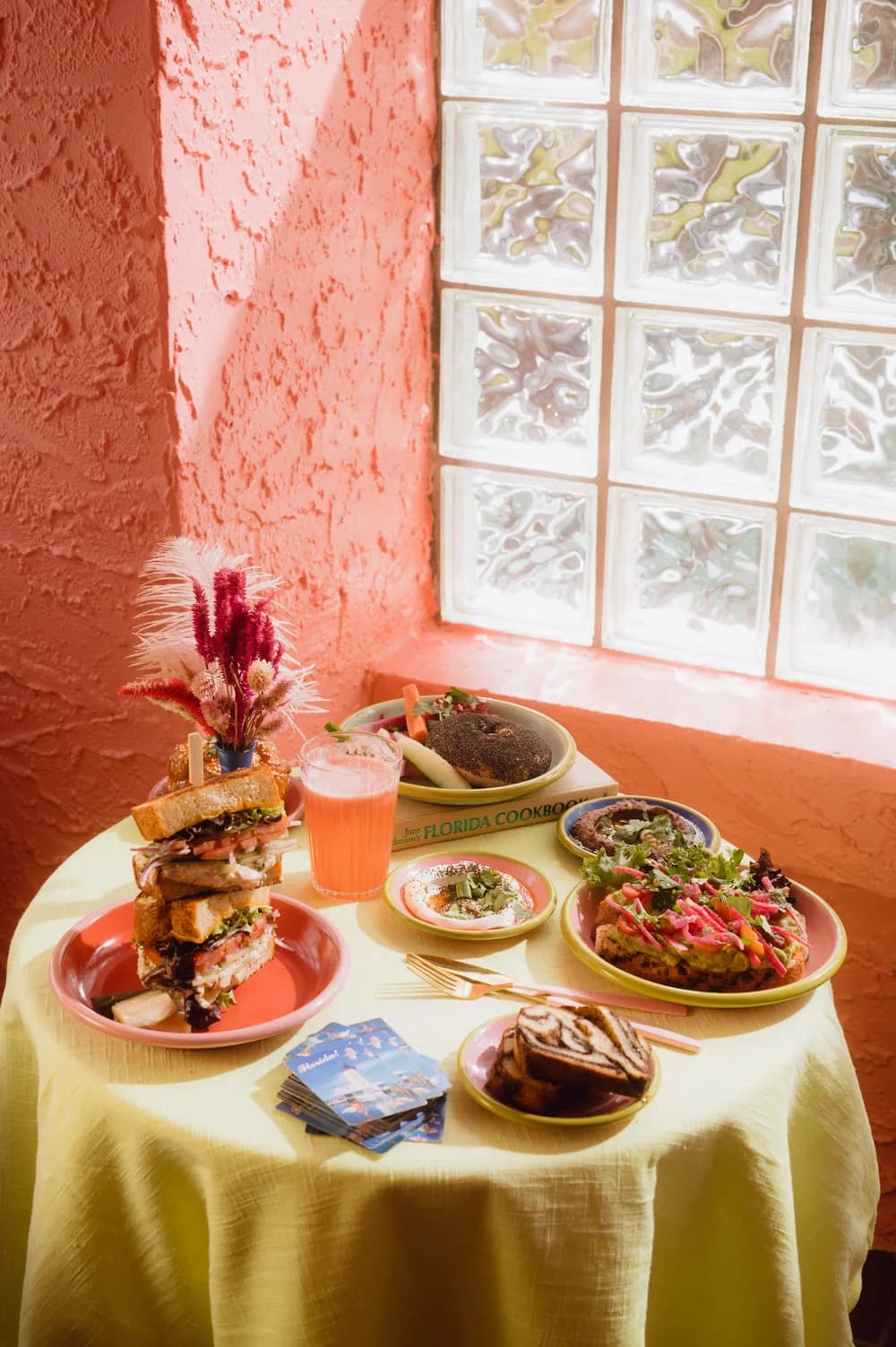 A spread at Potchke — a tall deli sandwich, a bagel, a babka slice, small plates, and a bright pink drink on a sunlit table