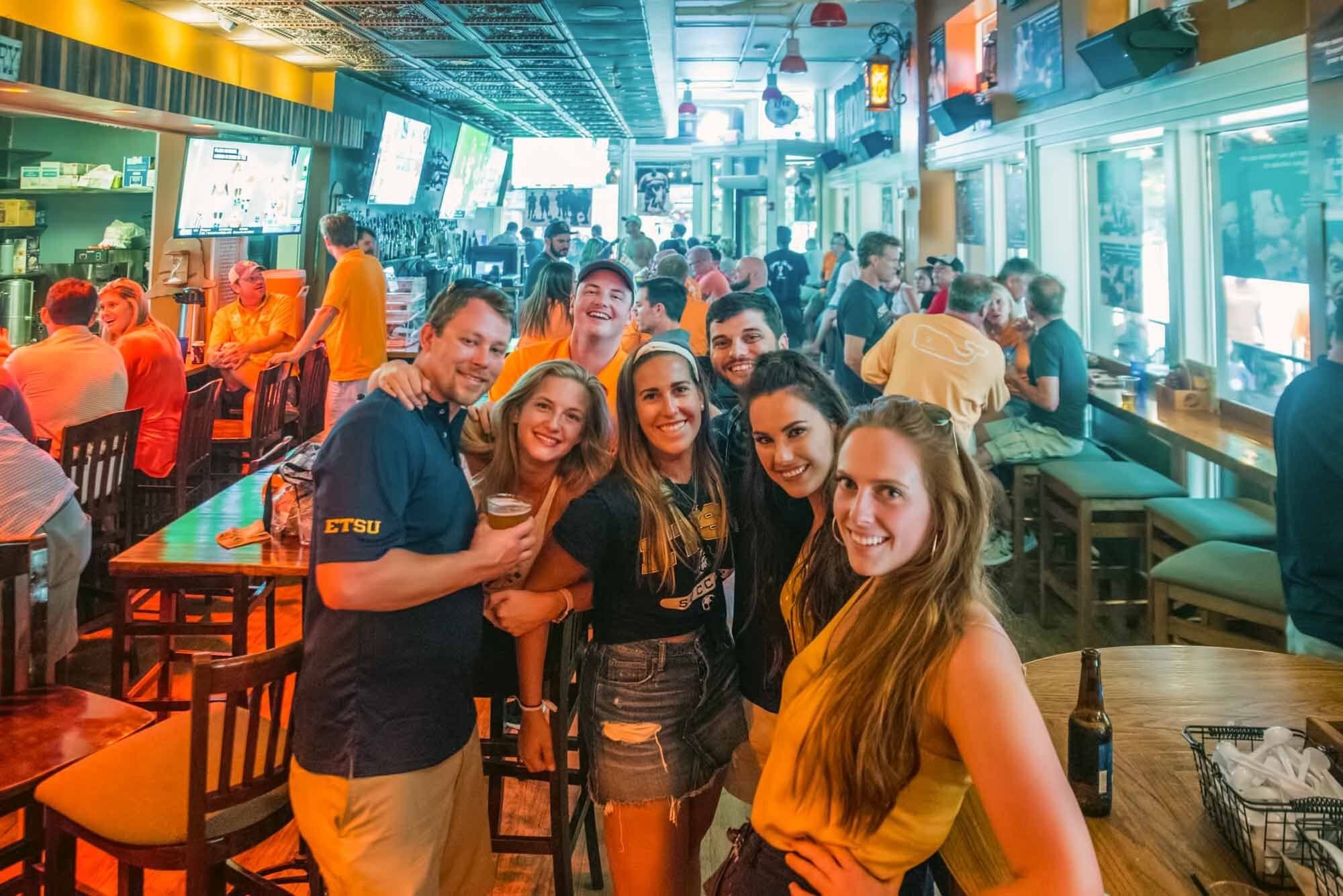 A group of fans posing together inside Tommy Trent's Sports Saloon on game day, TVs and a packed bar behind them