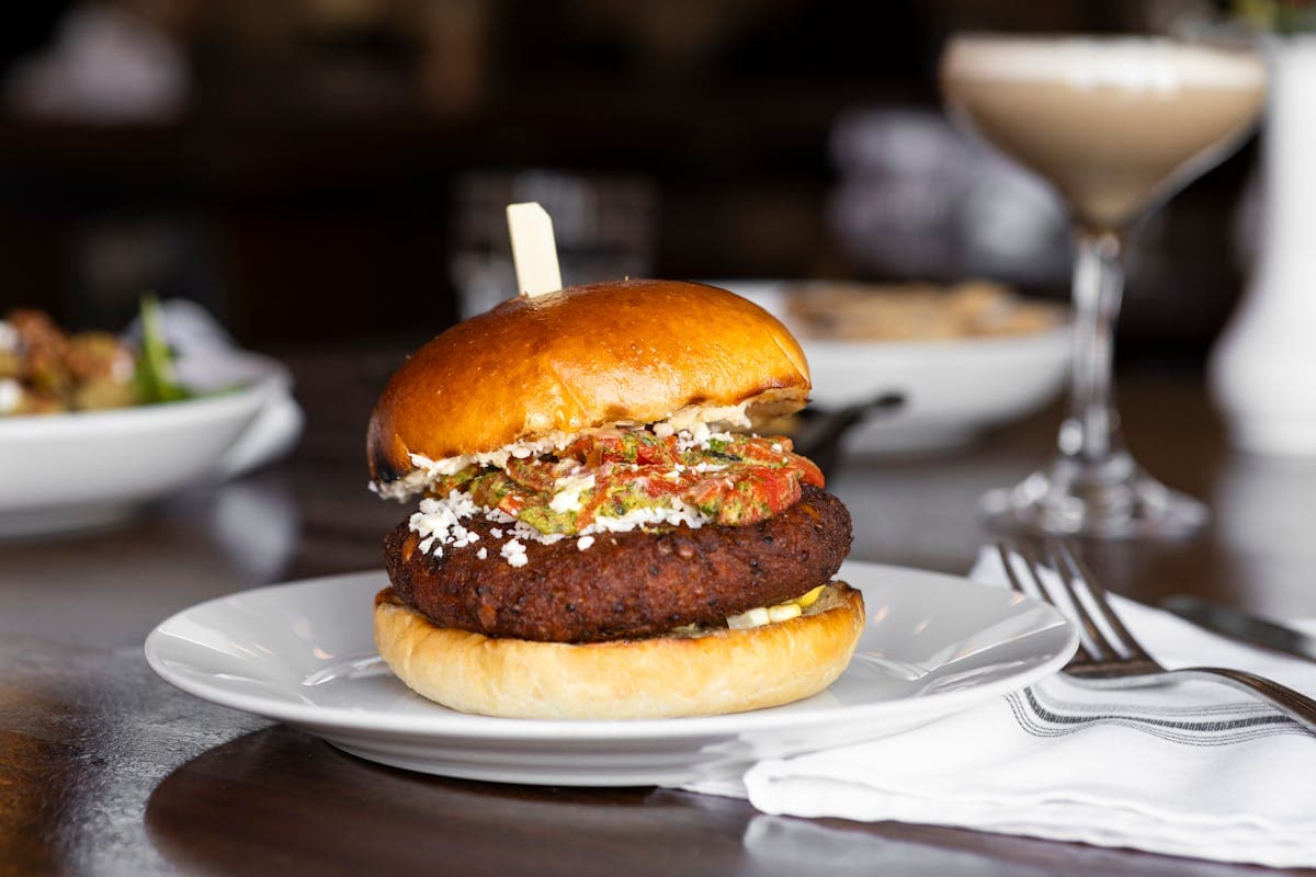 A burger topped with feta and roasted red pepper relish on a white plate, with a creamy cocktail in the background