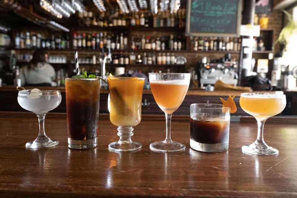A lineup of six colorful cocktails on The Bourgeois Pig's wood bar, backlit by rows of bottles and an espresso machine