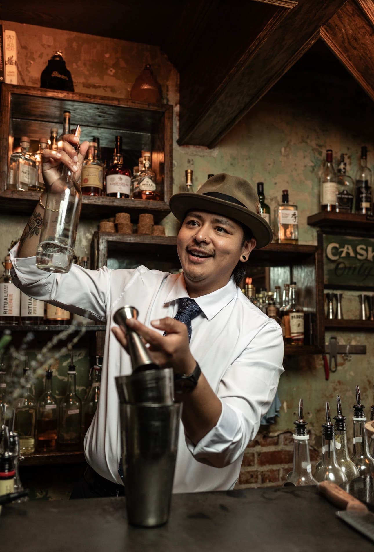 A bartender at The Other Room in a fedora and tie reaching for a bottle behind the bar with a cocktail shaker in hand