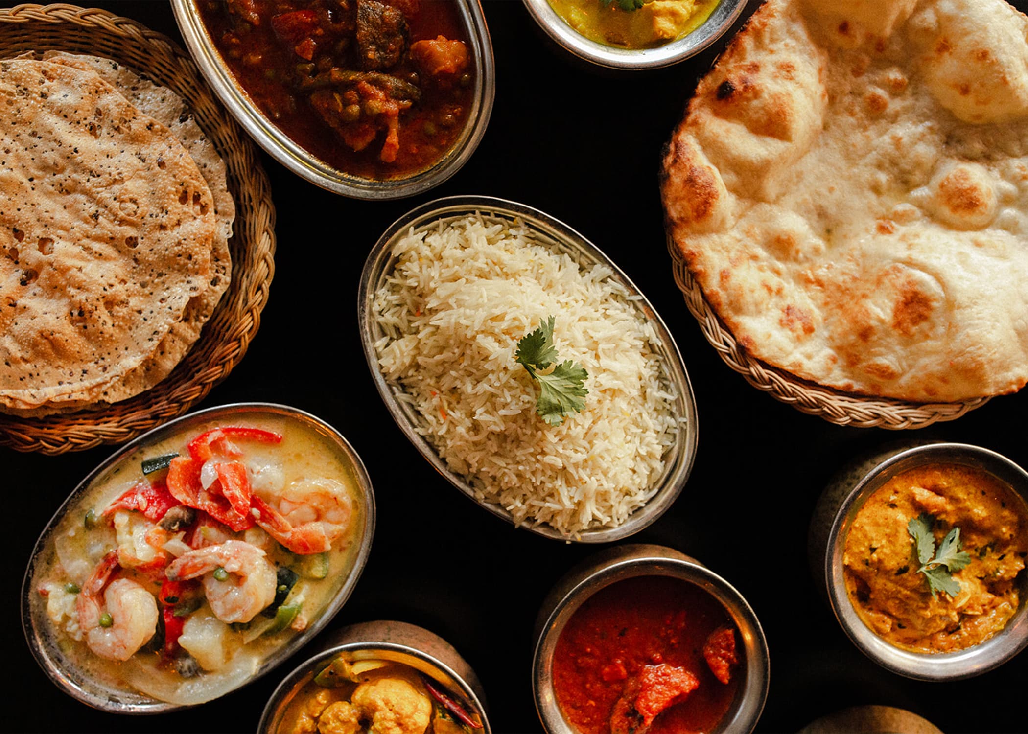 An overhead spread from The Oven — naan, basmati rice, and several curry and shrimp dishes in steel bowls