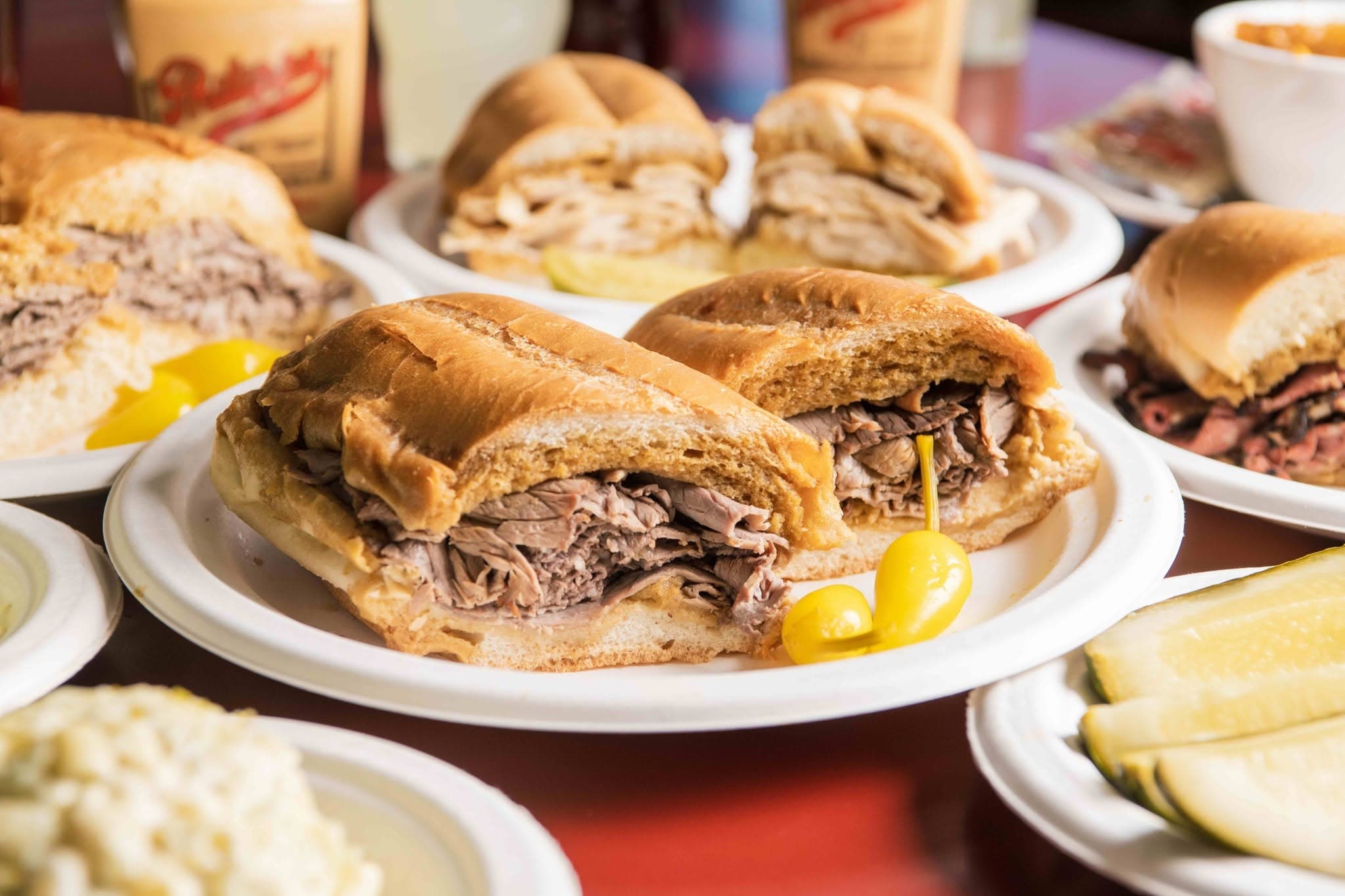 Halved French dip sandwiches piled with sliced beef on plates, with pickled peppers and sides on the table
