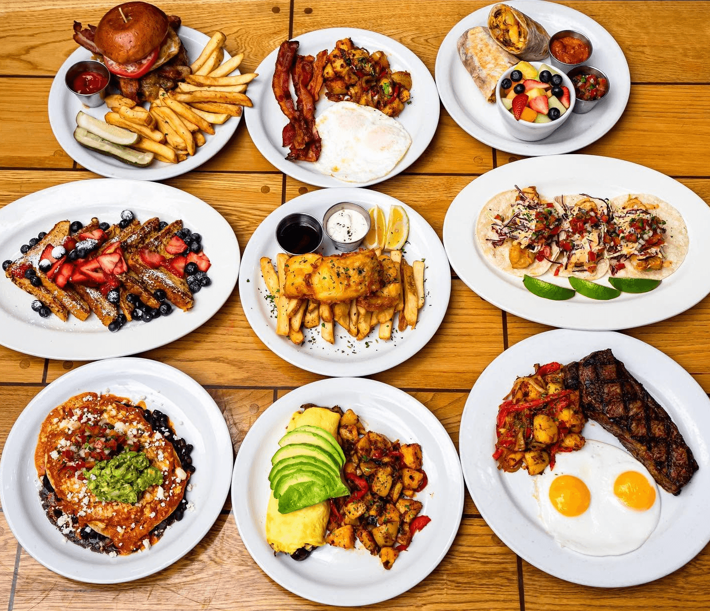 Overhead spread of nine brunch plates from The Galley — burgers, fish and chips, fish tacos, huevos, and steak and eggs on a wood table