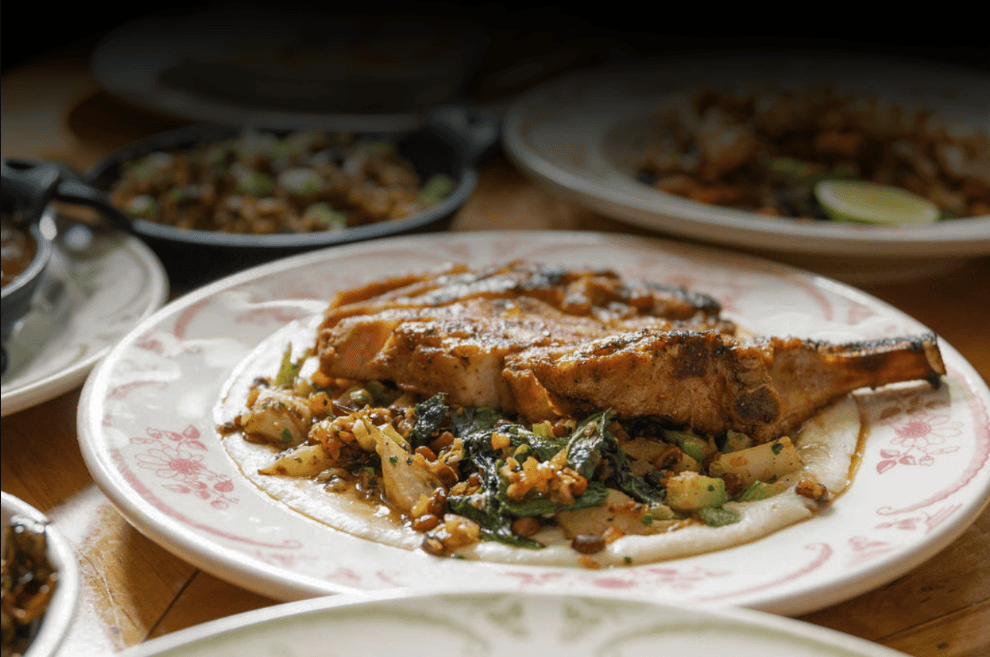 A seared bone-in pork chop plated over greens and grits with pecans at North of Bourbon