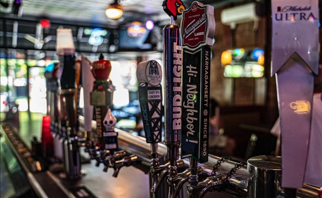 Beer taps lined up at The Granville Pub's bar, including a Louisville Cardinals-branded handle, with the dim game-day room behind