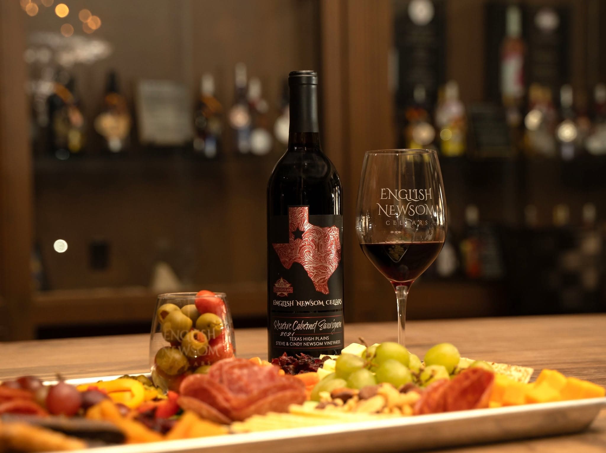 A bottle and glass of English Newsom Cellars Reserve Cabernet Sauvignon beside a charcuterie board of meats, cheeses, olives, and grapes