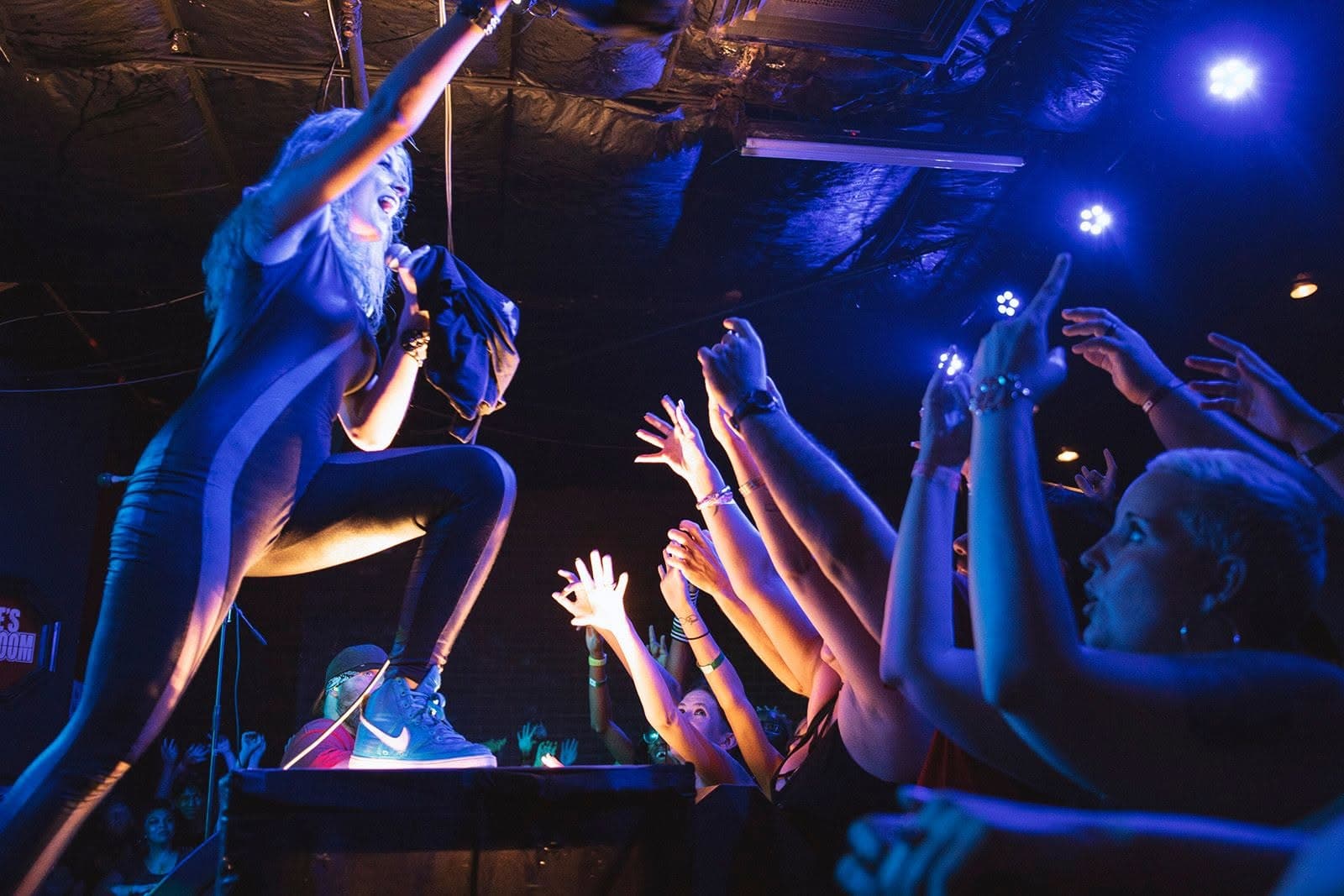 A performer on stage reaching toward a crowd with raised hands under blue stage lights inside Jake's Backroom