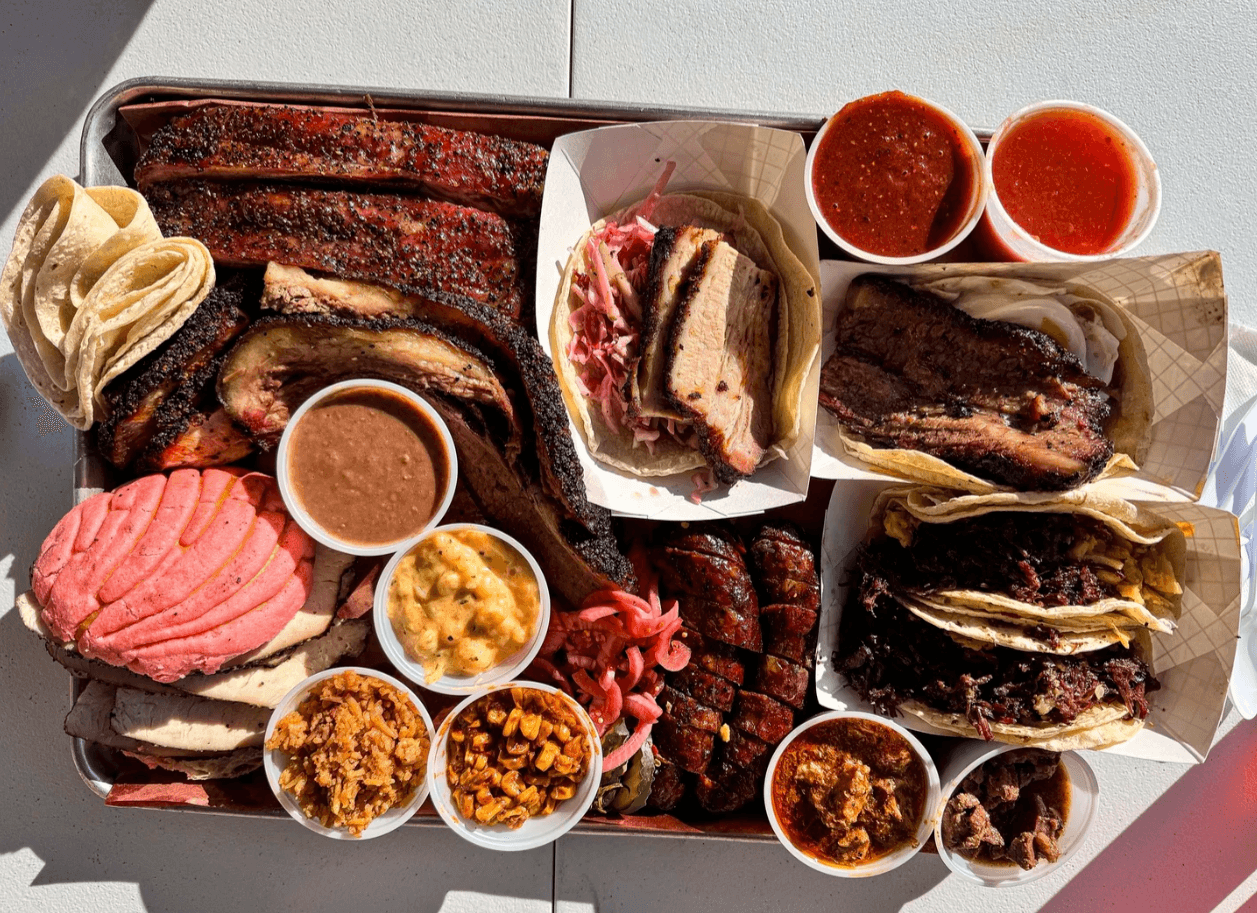 Overhead spread of a full barbecue platter — ribs, brisket, sliced tacos, sausage, salsas, and a pink concha, plus sides