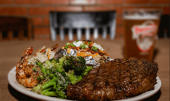 A hand-cut ribeye plated with grilled shrimp, broccoli, and a loaded baked potato at Triple J Chophouse & Brew Co.