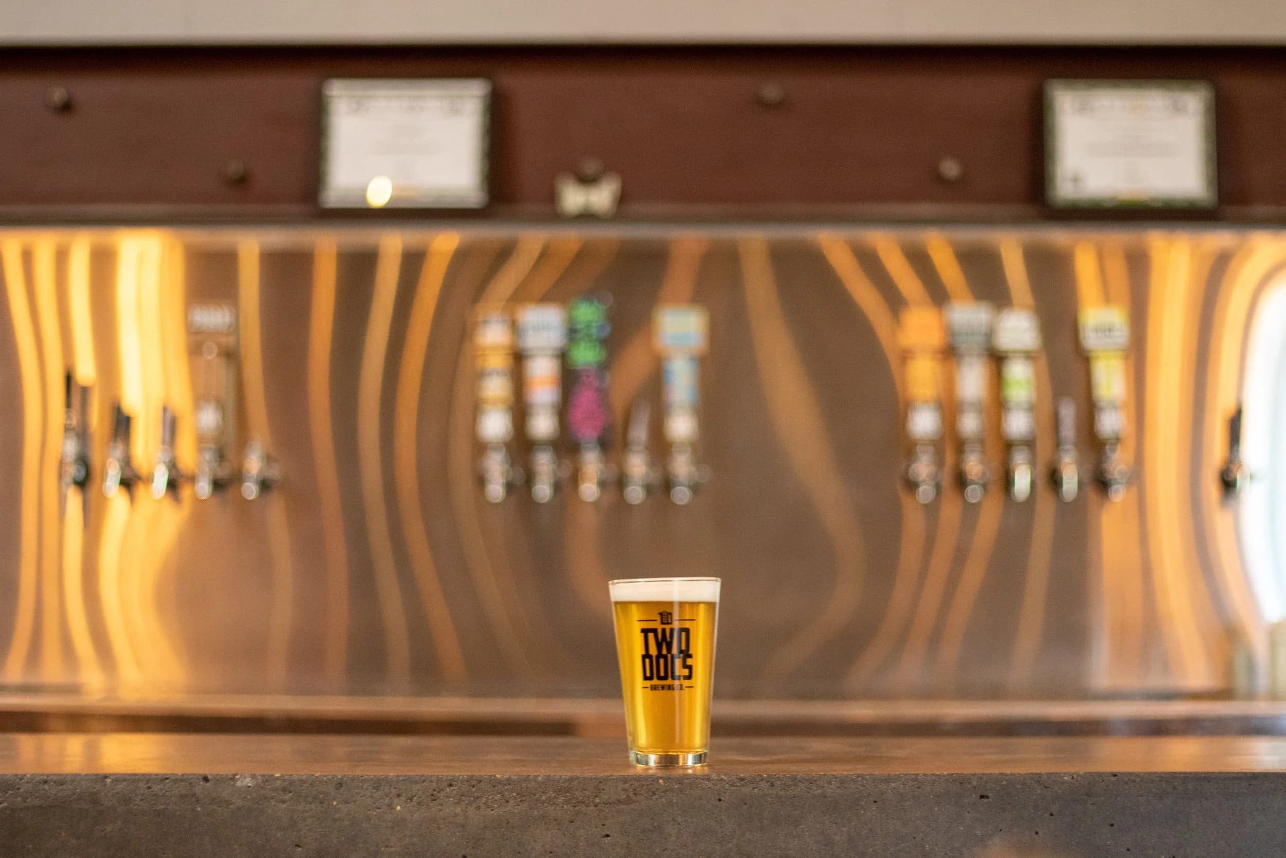 A branded Two Docs Brewing Co. pint glass of golden beer on the bar with the row of tap handles behind it