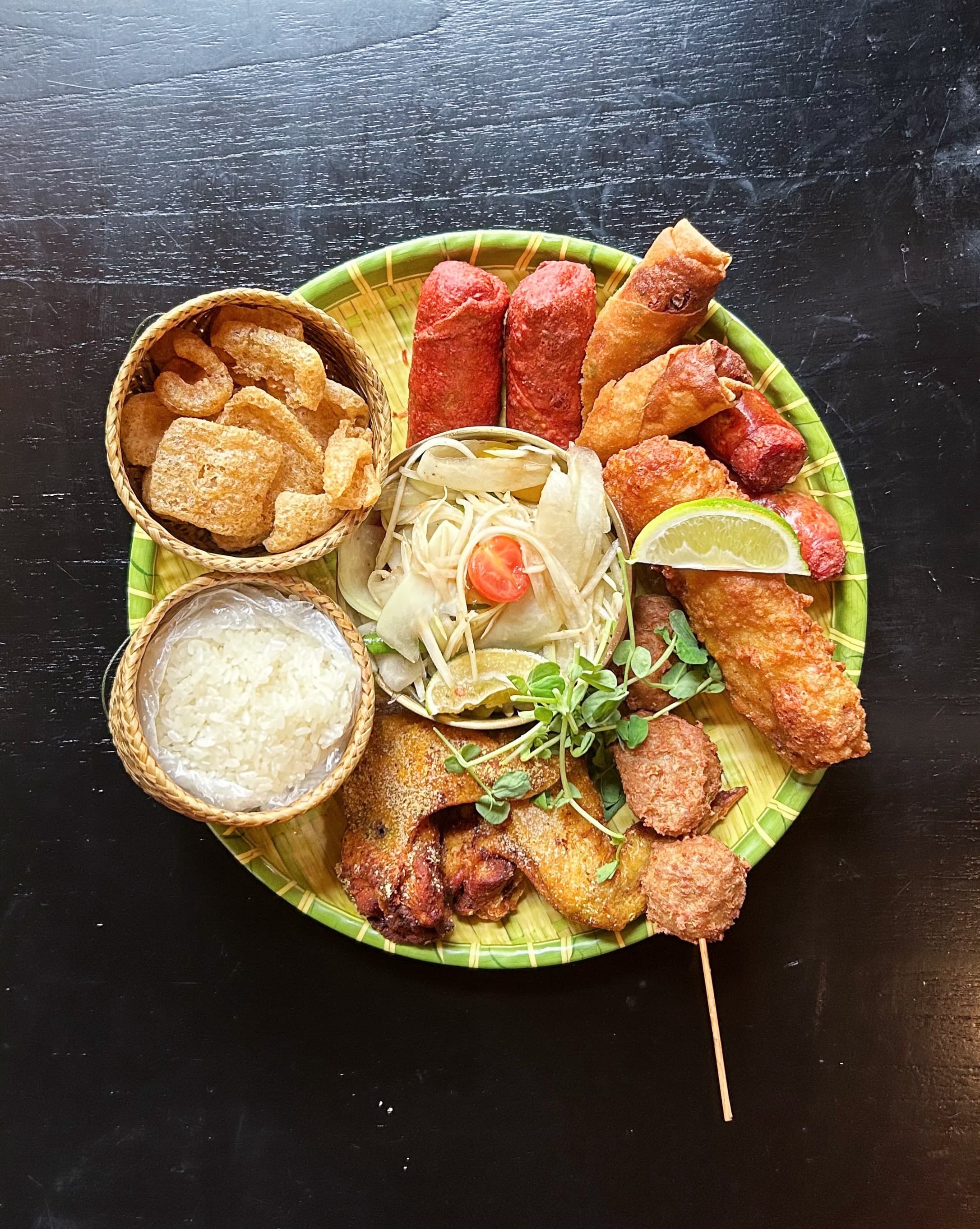 Overhead shot of a Laotian sampler platter — fried chicken wings, sai oua sausage, spring rolls, papaya salad, sticky rice, and pork rinds
