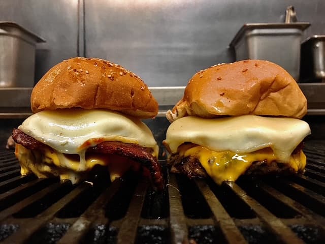 Two double cheeseburgers with bacon on the grill at Dotty Dumpling's Dowry, cheese melting over the patties