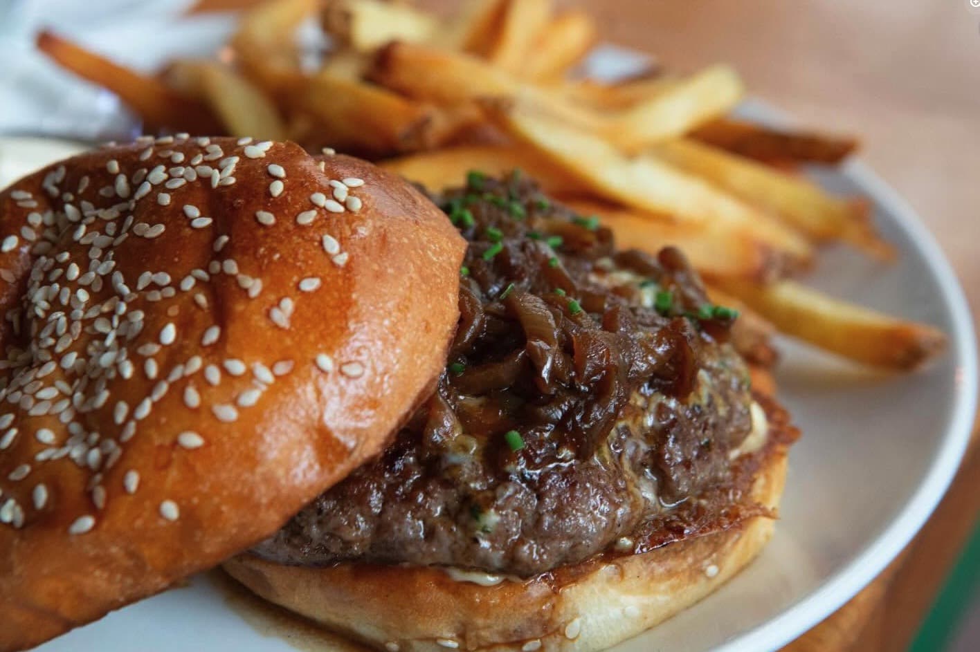 Close-up of a burger with caramelized onions and chives on a sesame bun, fries in the background