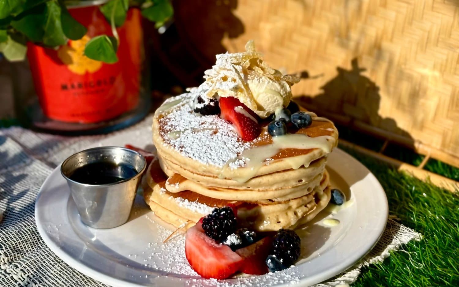 A stack of pancakes topped with whipped cream, strawberries, blackberries, blueberries, and powdered sugar, with maple syrup on the side, at Marigold Kitchen