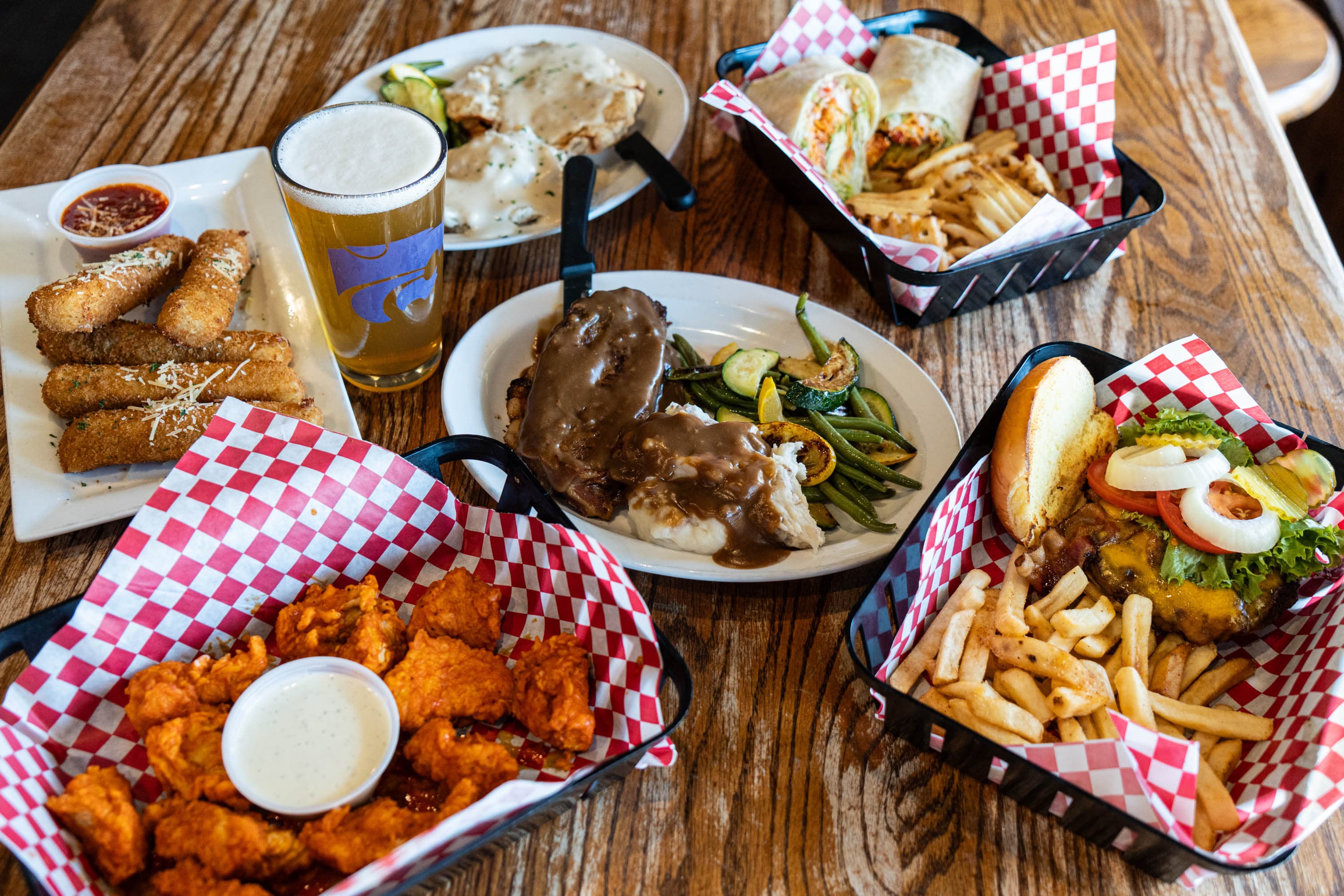 A loaded table at Kite's Bar & Grill with wings, mozzarella sticks, a wrap and fries, chicken-fried steak, and a K-State pint of beer