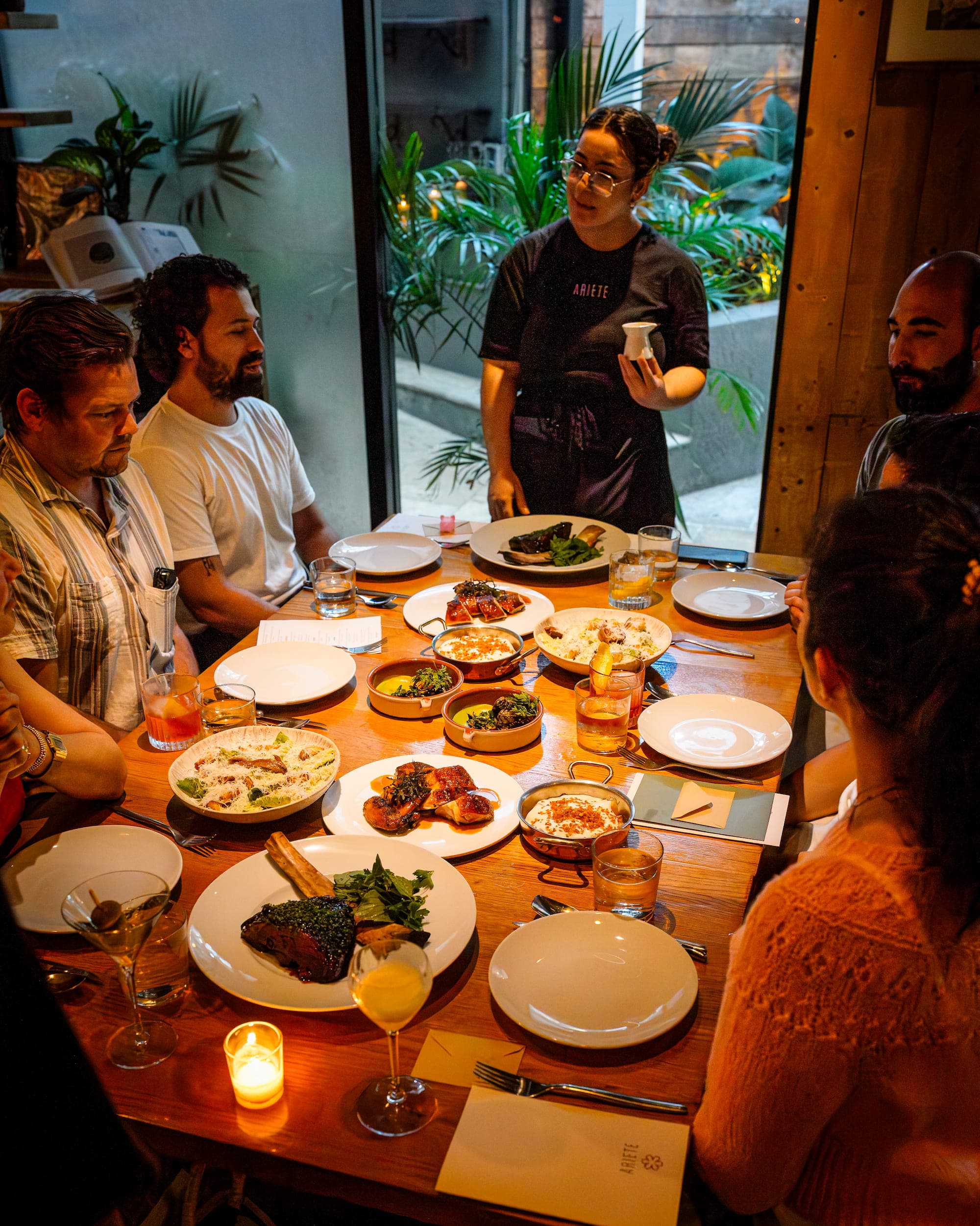 A shared table at Ariete with a chef presenting a dish to a group of diners, roast chicken, salads, and cocktails spread across the table