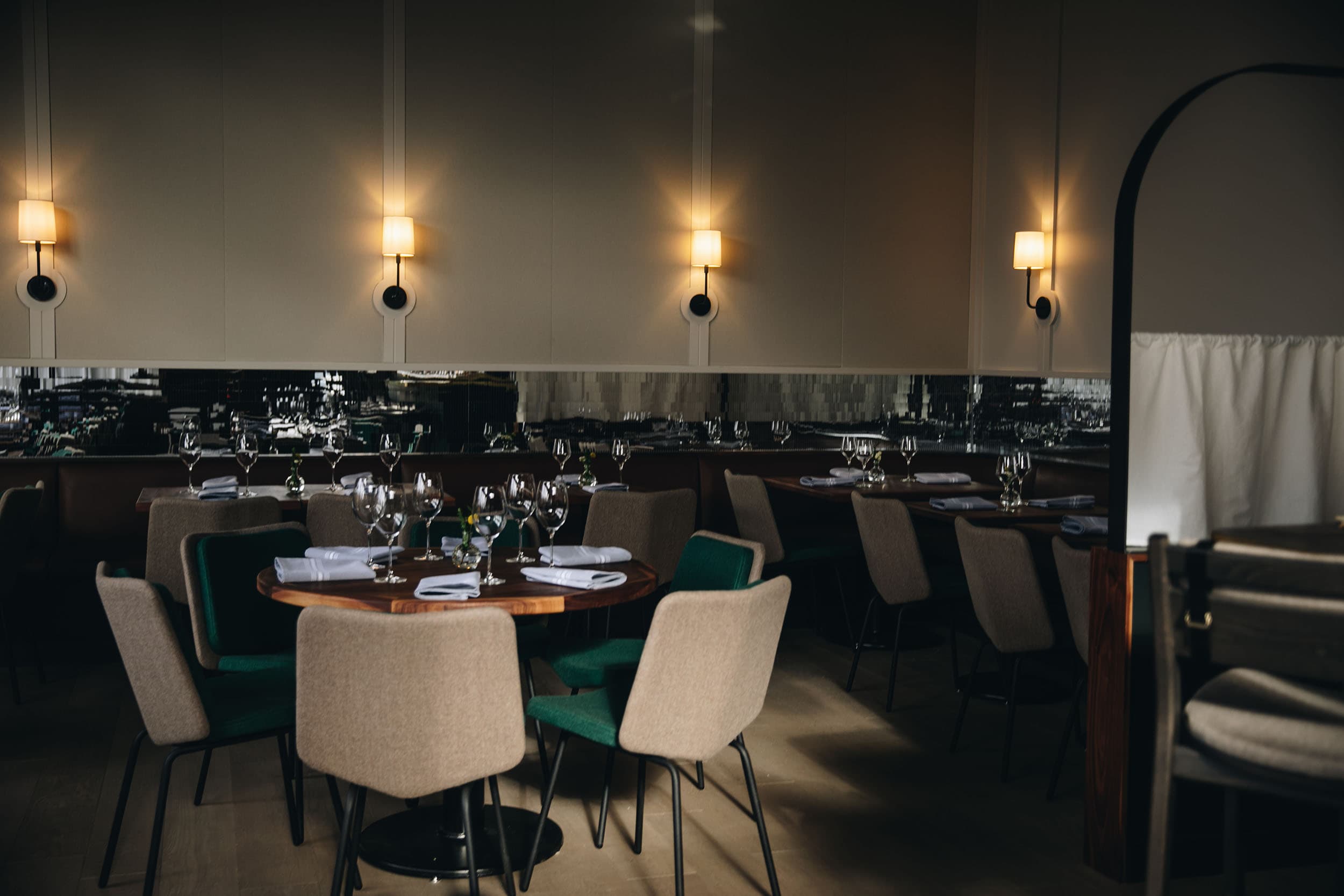 Bûcheron's warm dining room with green velvet chairs, wine glasses set on a round table below a mirrored bar backdrop