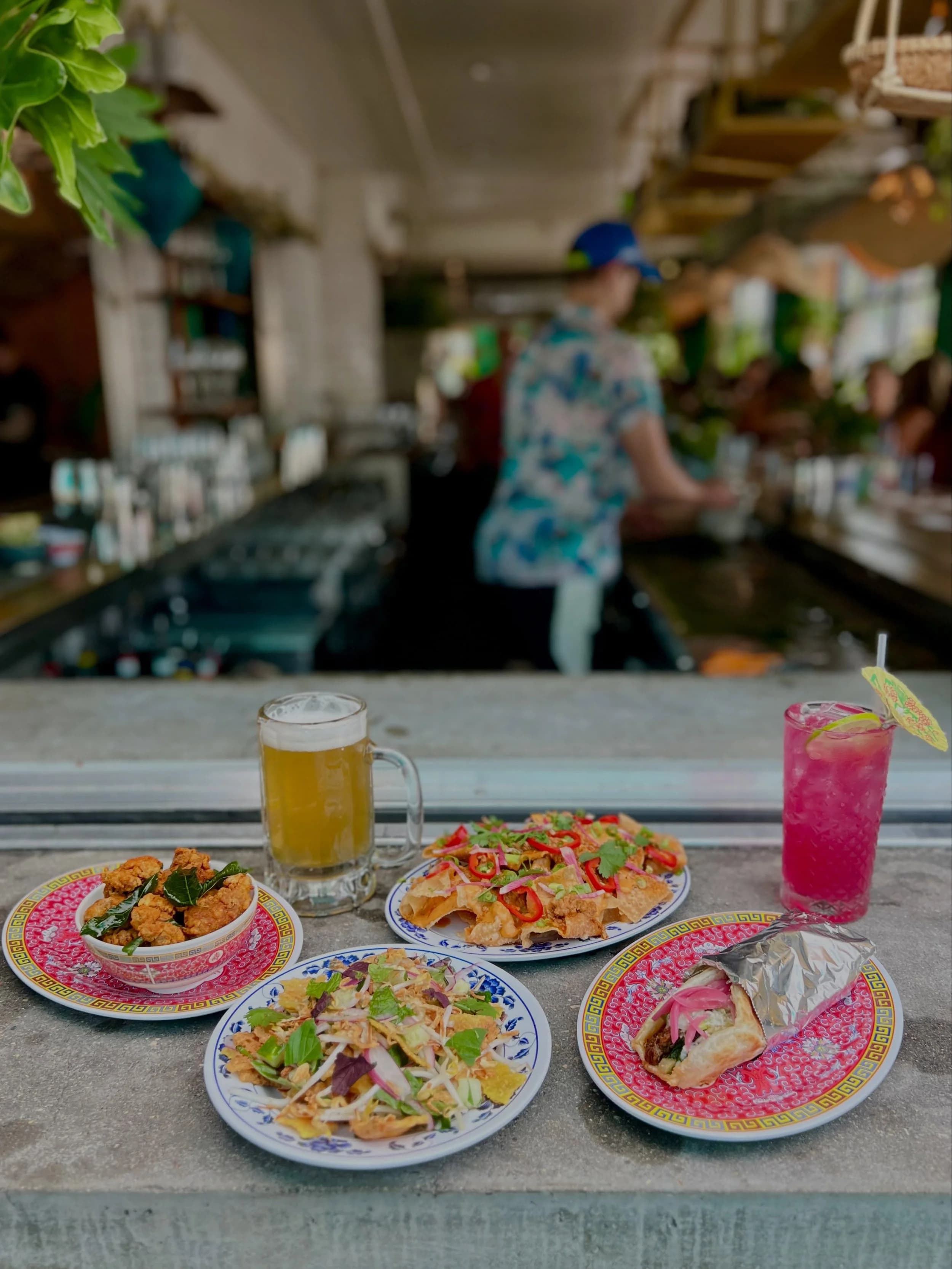 A spread of Hai Hai dishes and cocktails on the bar rail — fried chicken, crispy rice salad, a beer, and a bright pink cocktail