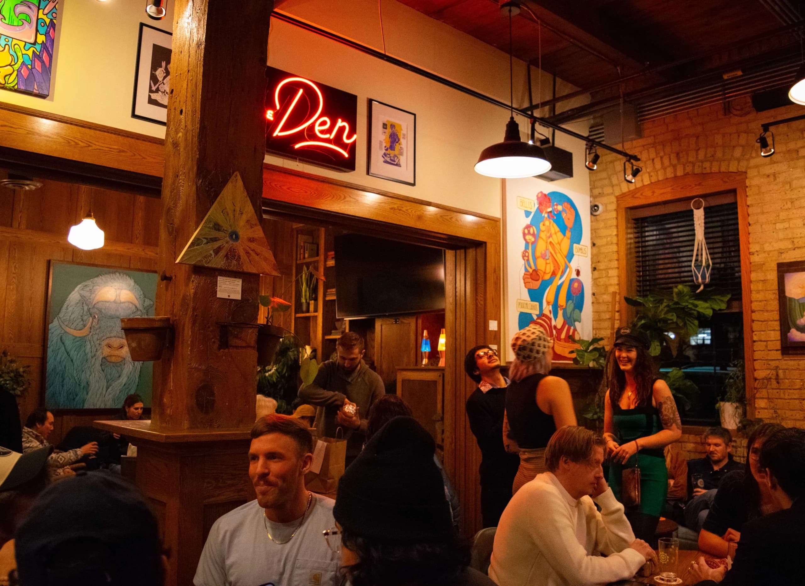 Indeed Brewing's taproom lounge area with a neon 'Den' sign, exposed brick, plants, and a crowd mingling under warm lighting