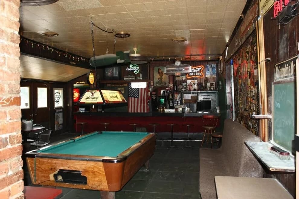 Springwater's dim dive-bar interior with a pool table, neon beer signs, and an American flag behind the bar