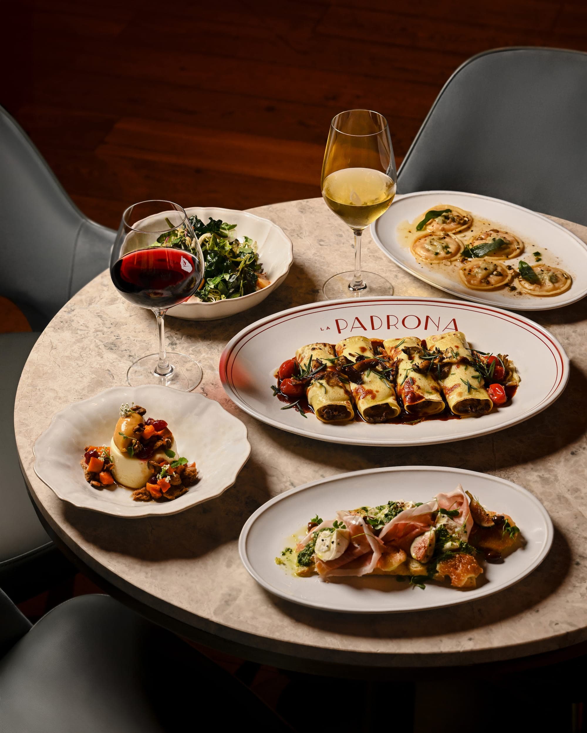 A table at La Padrona set with a branded platter of filled pasta, ravioli, a burrata dish, wine, and a salad