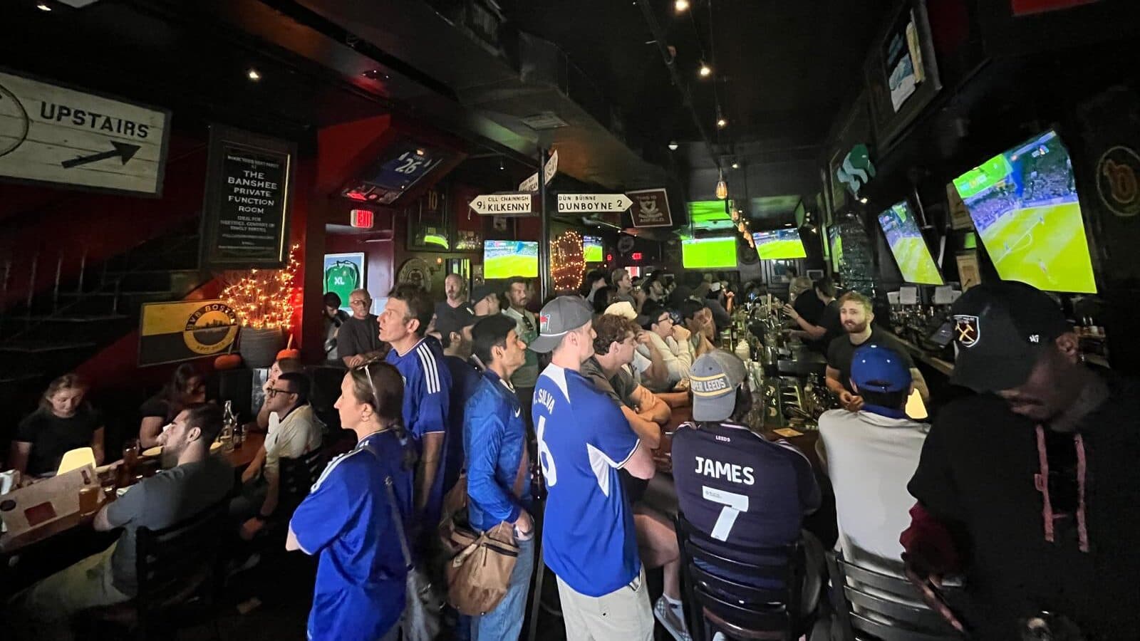 A packed crowd inside The Banshee watching a soccer match on the wall of screens, jerseys and pints in hand