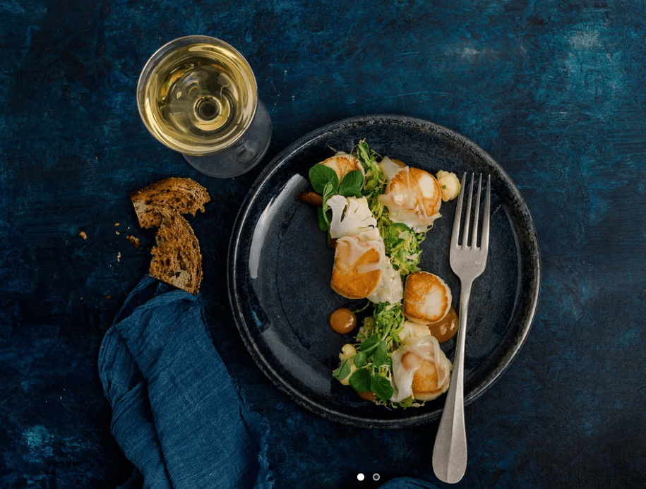 Seared scallops with pea shoots and cauliflower on a dark plate, a glass of white wine and torn bread alongside