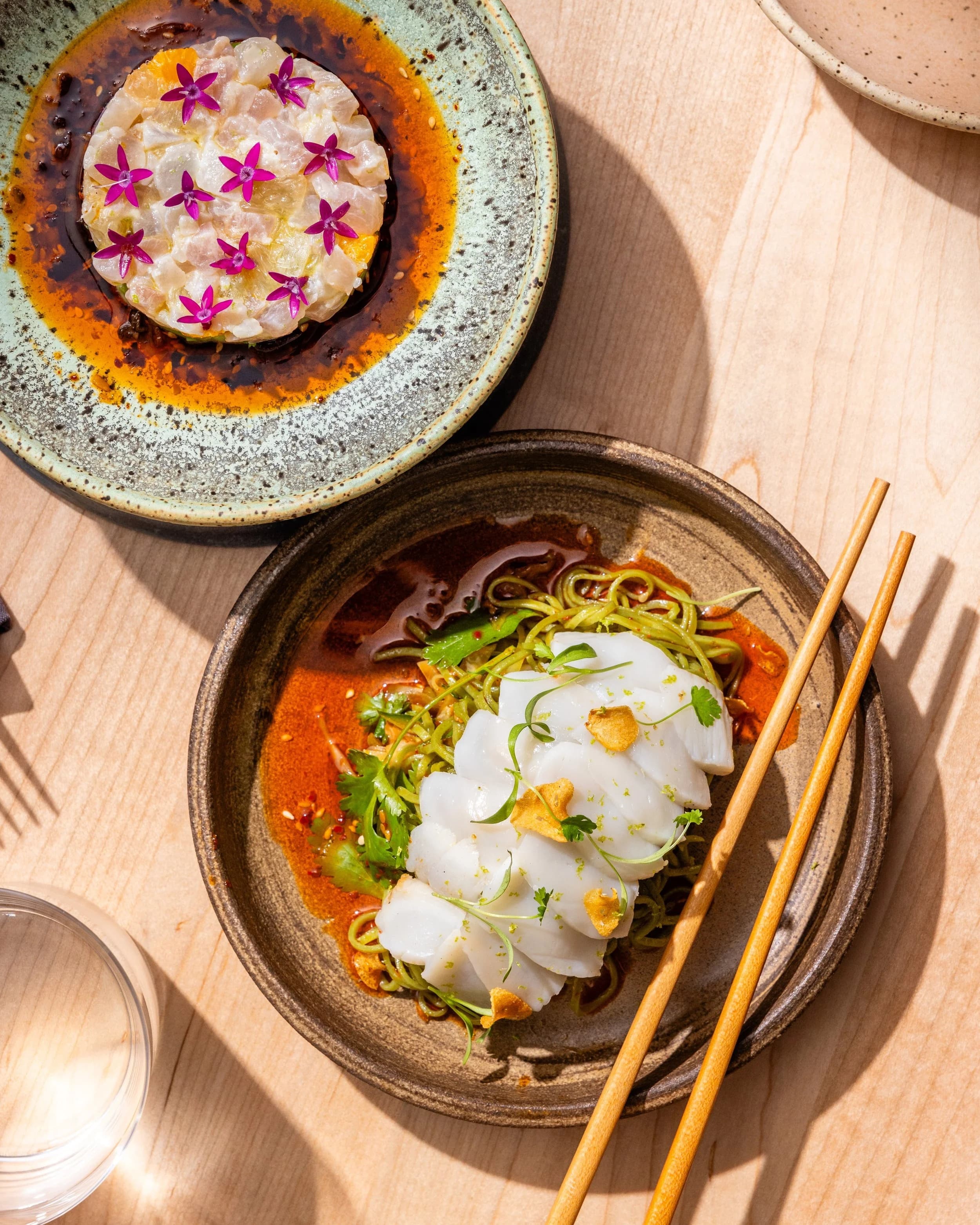Two artfully plated small dishes at Coro — a citrus crudo topped with edible flowers, and sliced raw fish over cold noodles with herbs