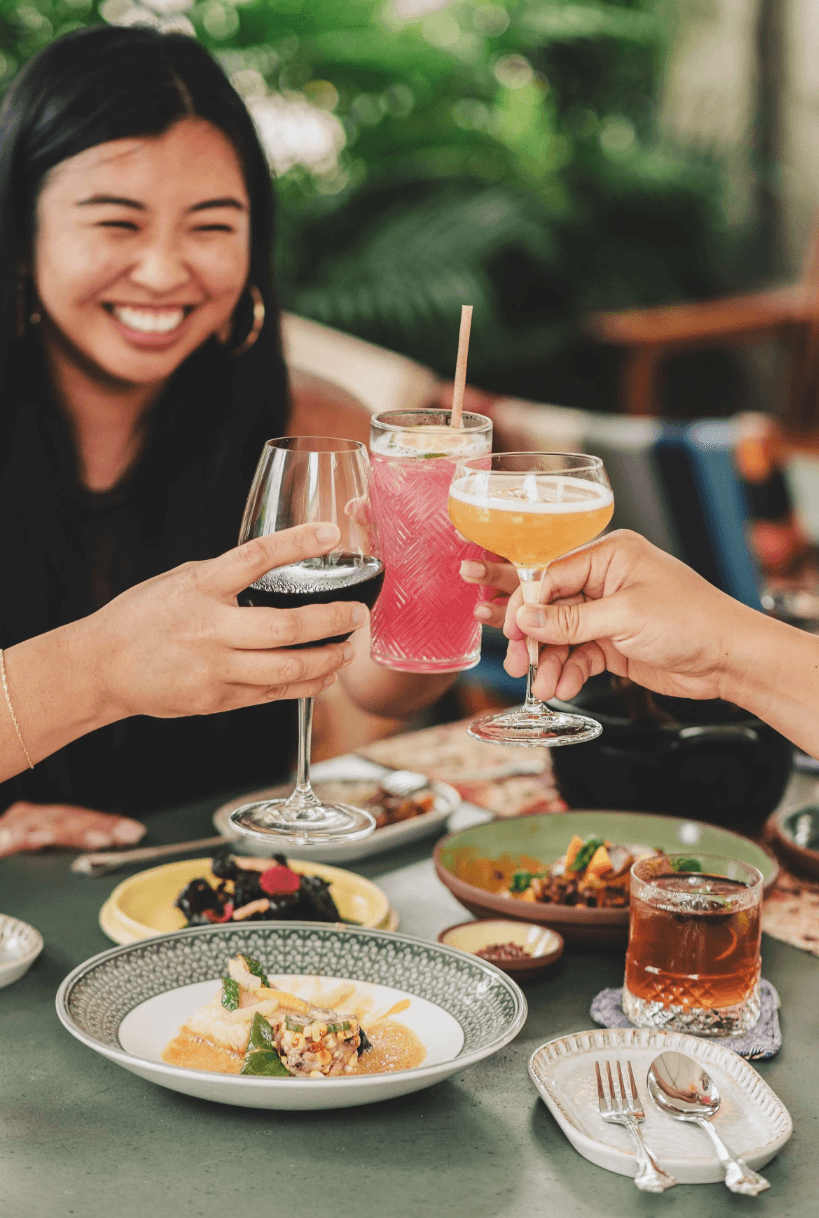 Friends toasting with wine and cocktails over a spread of shared plates on Kaya's leafy patio