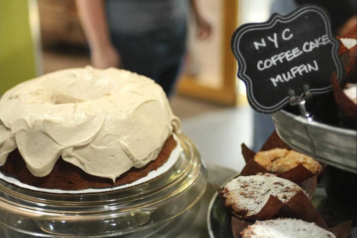 A frosted layer cake and powdered-sugar muffins on display in Se7en Bites' bakery case, with a chalkboard sign for the NYC Coffee Cake Muffin