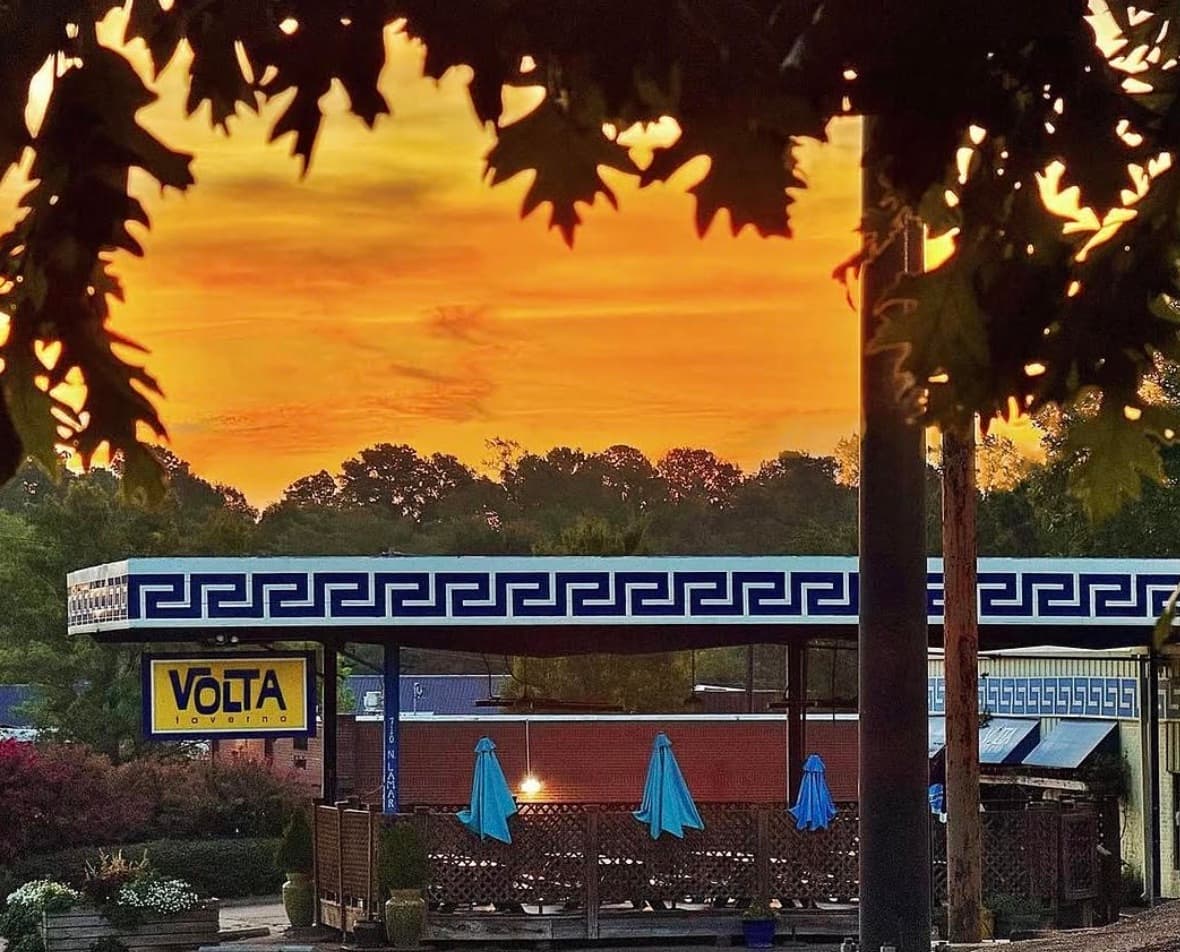 Volta Taverna's Greek-key roofline and patio umbrellas silhouetted against an orange sunset