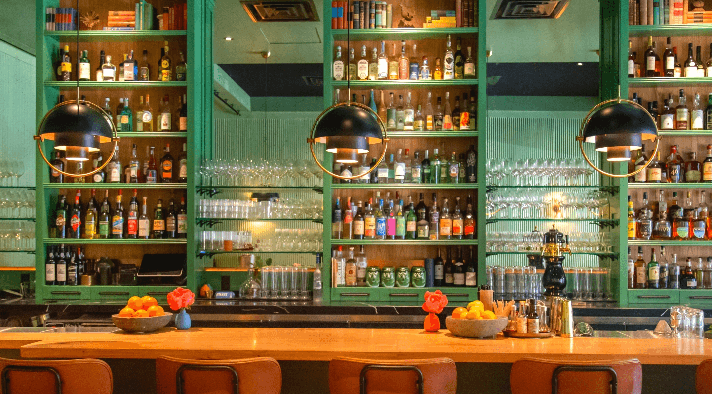 BarZola's green-shelved back bar lined with liquor bottles under globe pendant lights, oranges and roses on the counter