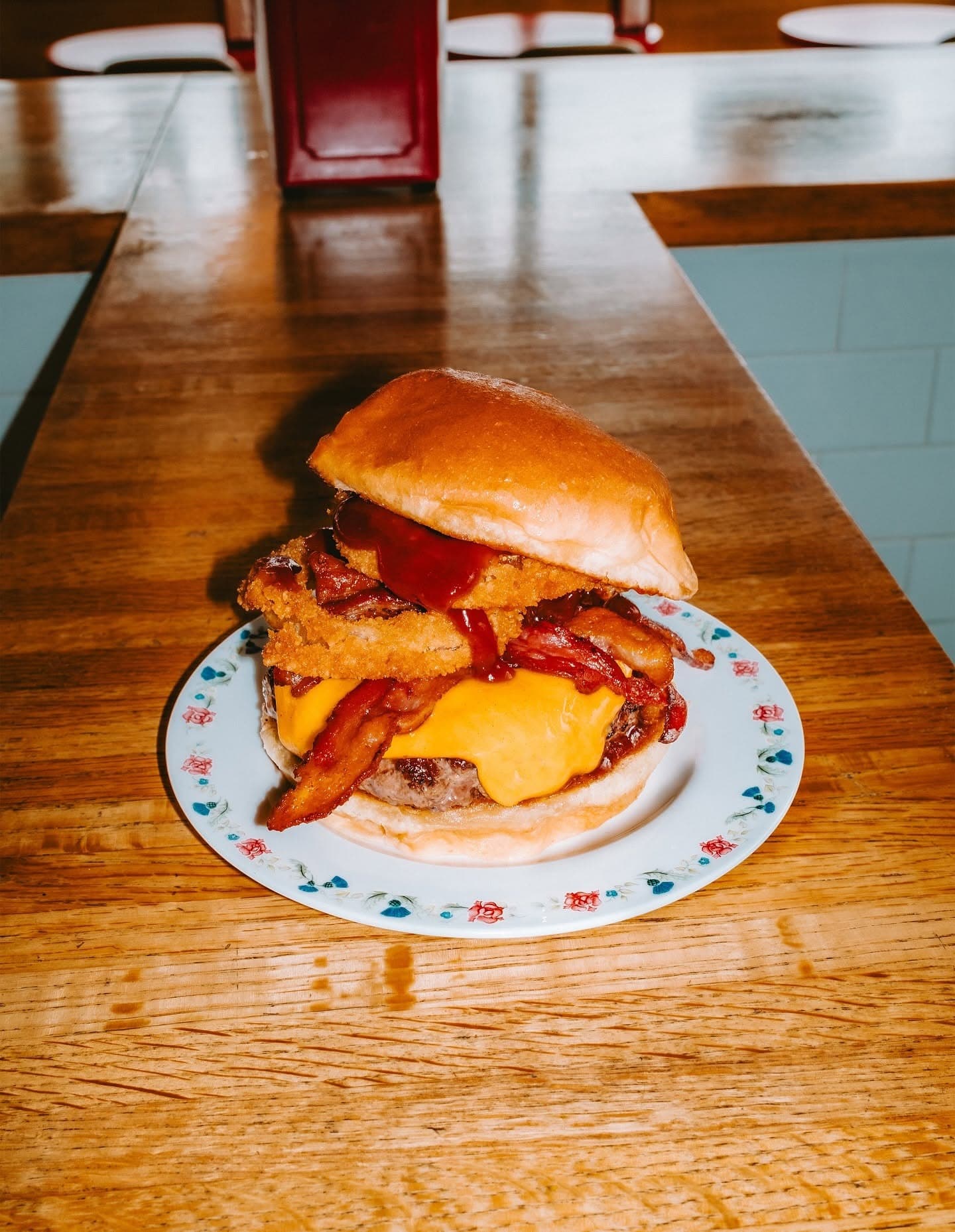 A cheeseburger stacked with bacon, an onion ring, and barbecue sauce on a floral-patterned plate at The Oinkster