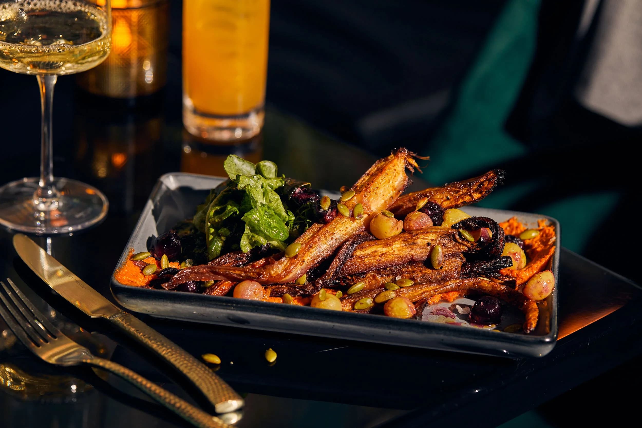 A plate of roasted carrots and root vegetables with pumpkin seeds and grapes over romesco, with a glass of wine and a cocktail on the table at Con Alma