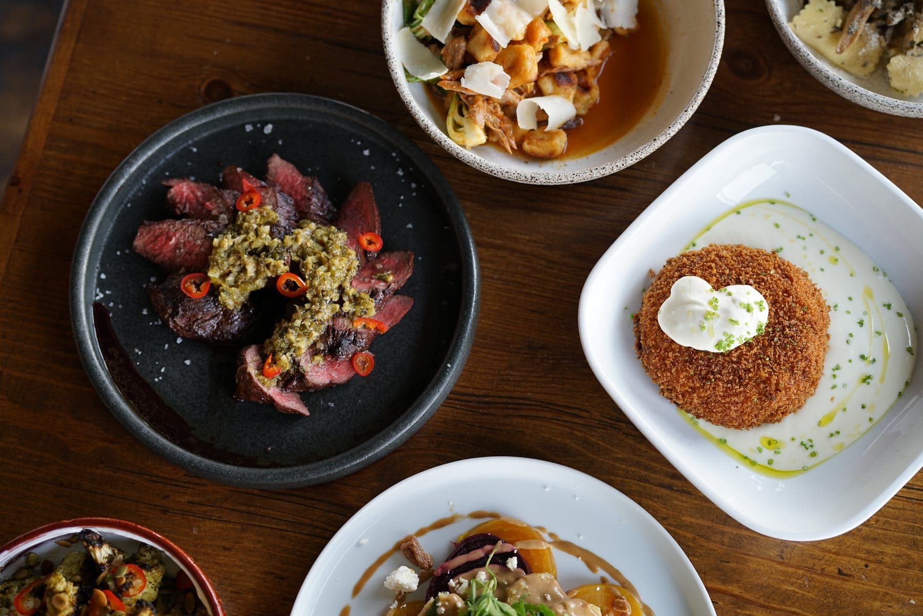 Small plates of steak with chimichurri, a crispy potato rösti, gnocchi and roasted vegetables plated at Communal