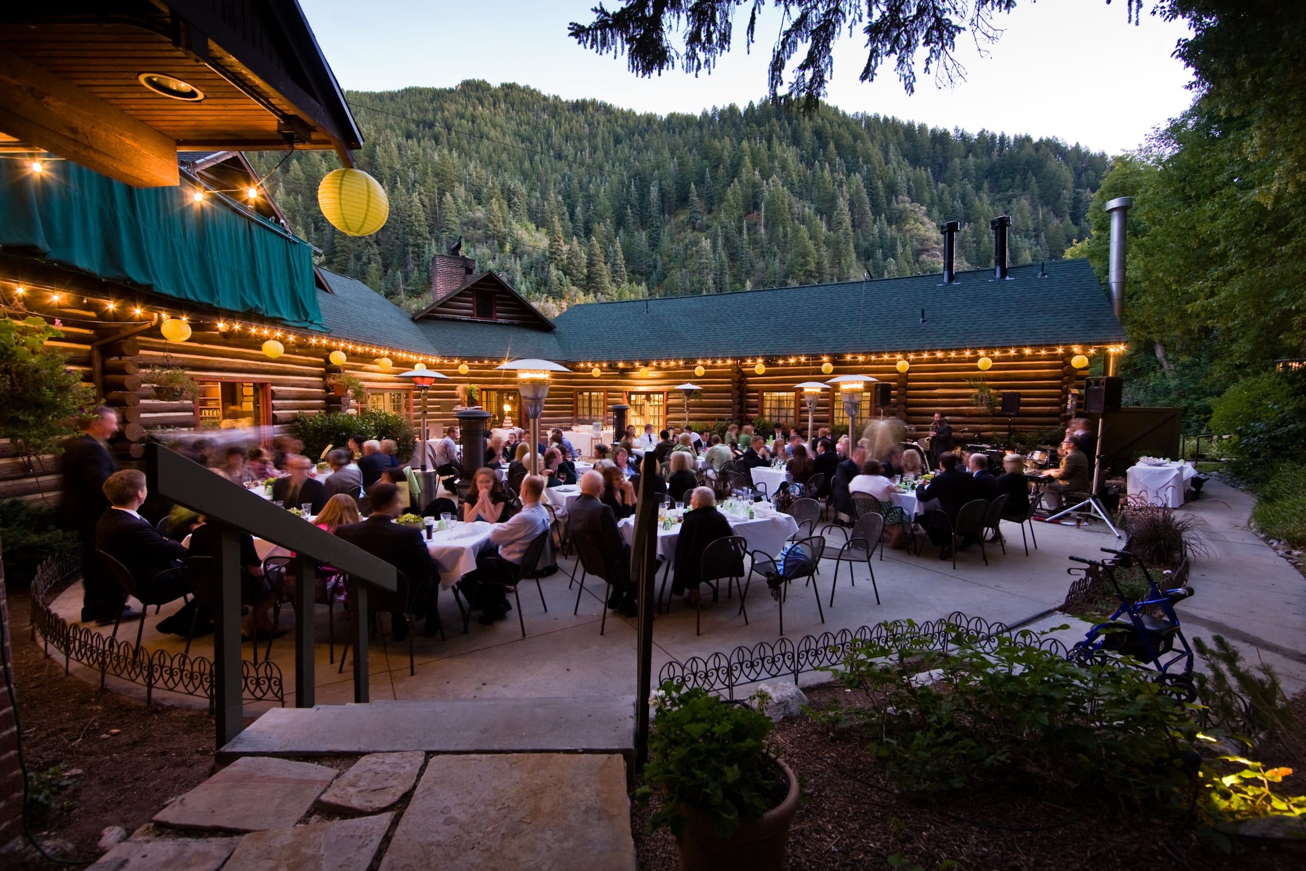 Guests dining at long tables on Log Haven's twilight patio, string lights and paper lanterns strung along the historic log building against a forested canyon backdrop