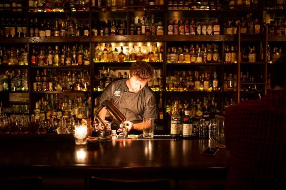 A bartender at Canon pouring a cocktail behind the bar, backed by floor-to-ceiling shelves of spirits under warm low light