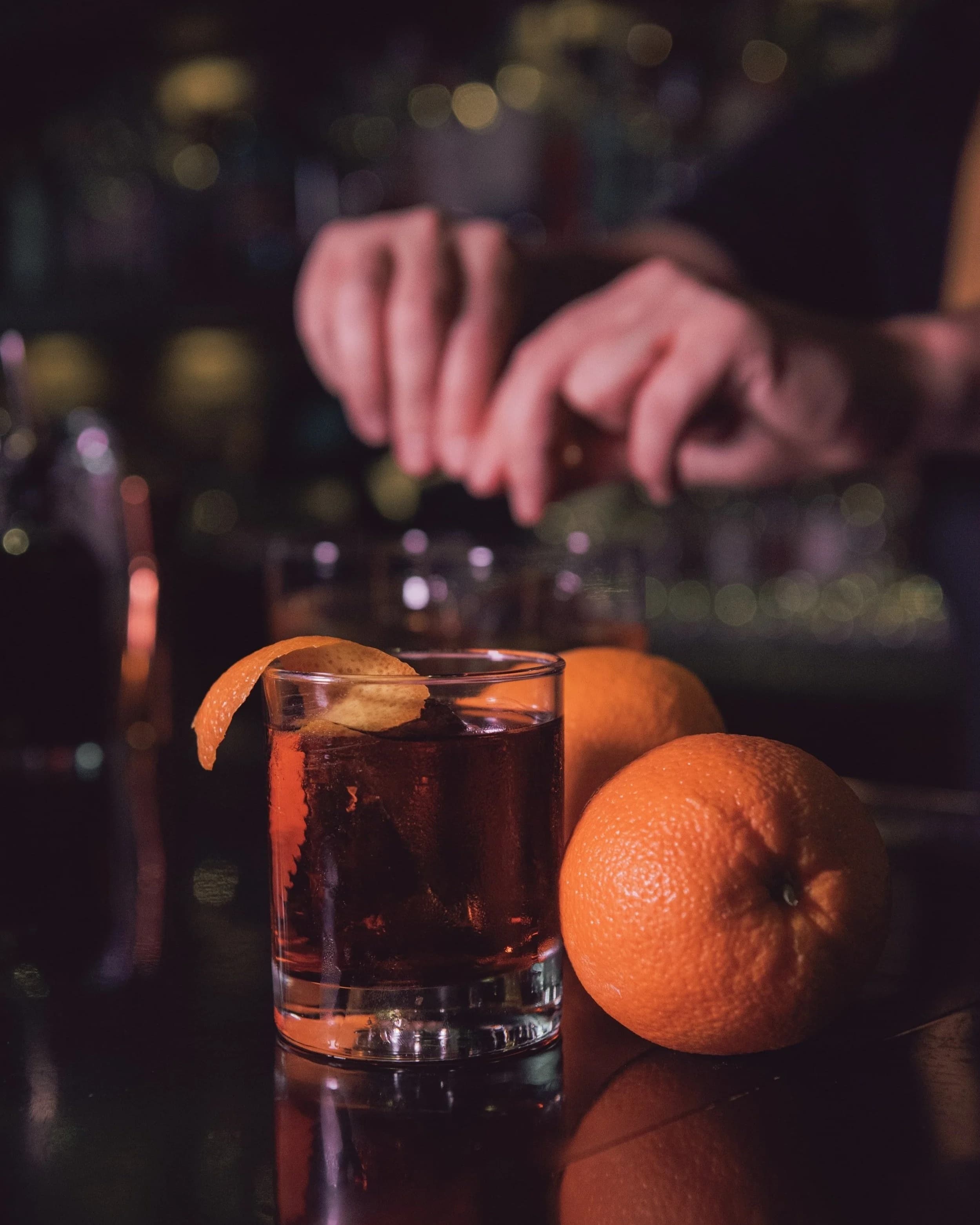 A dark spirit cocktail garnished with an orange twist on the bar at Rob Roy, with a bartender working behind it and fresh oranges nearby