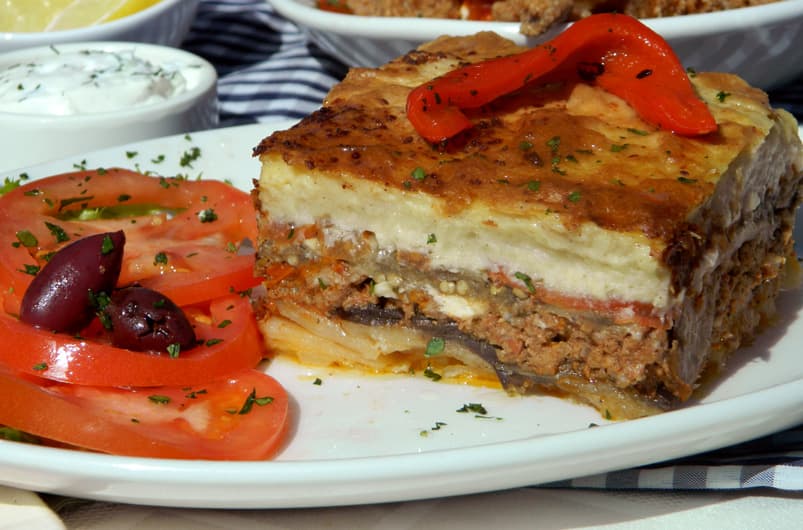 A slice of layered moussaka topped with red pepper next to a Greek tomato and olive salad