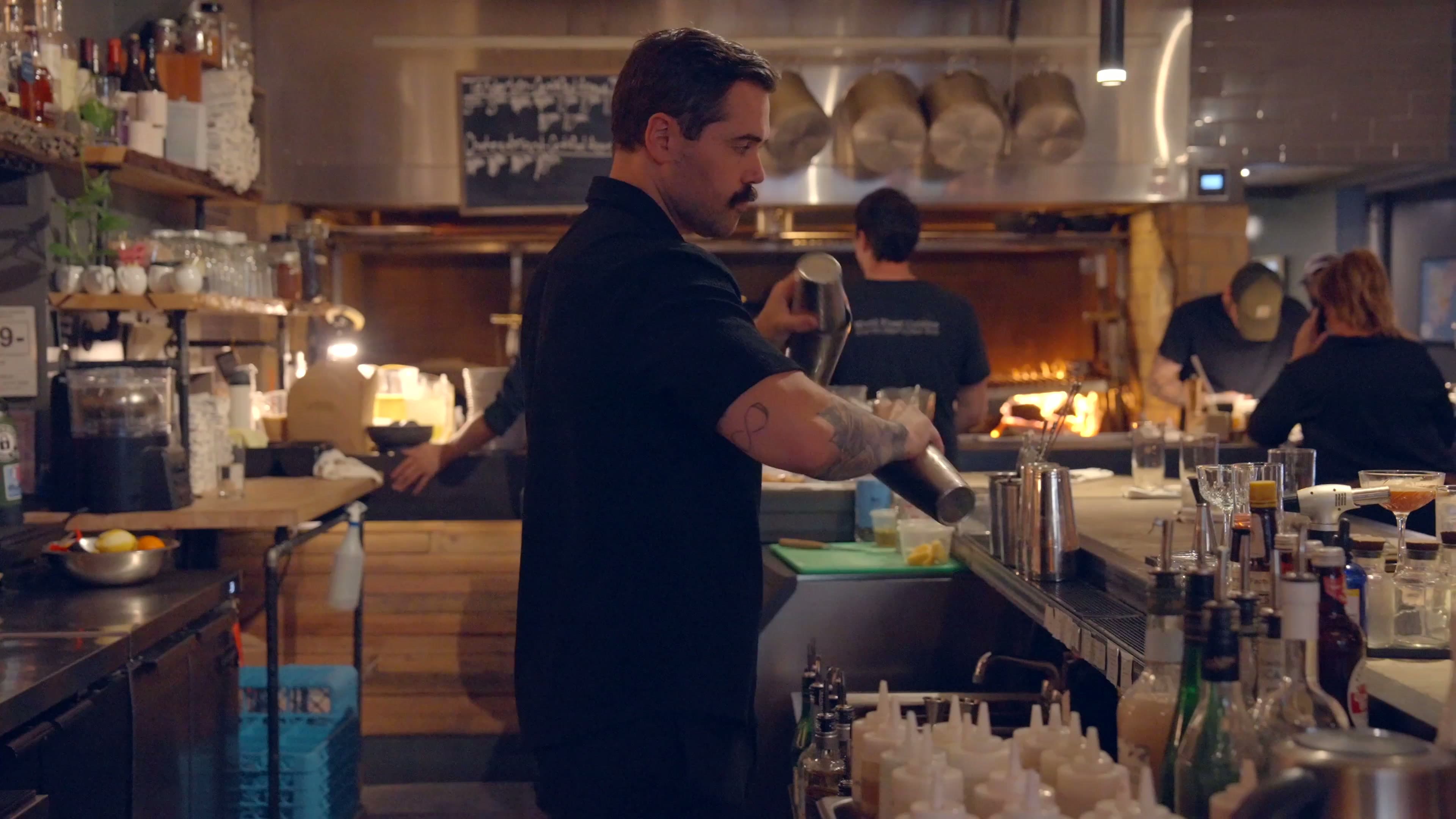 A bartender at Eden shaking a cocktail with the open kitchen's flames visible behind