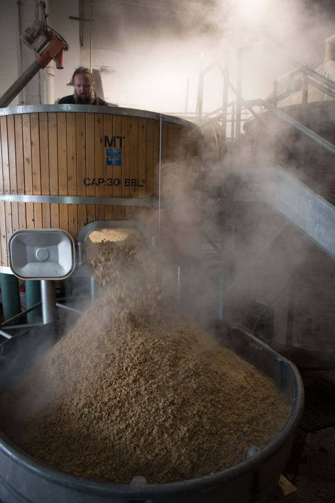 A brewer emptying steaming spent grain from a mash tun into a vessel at Middle Ages Brewing Company