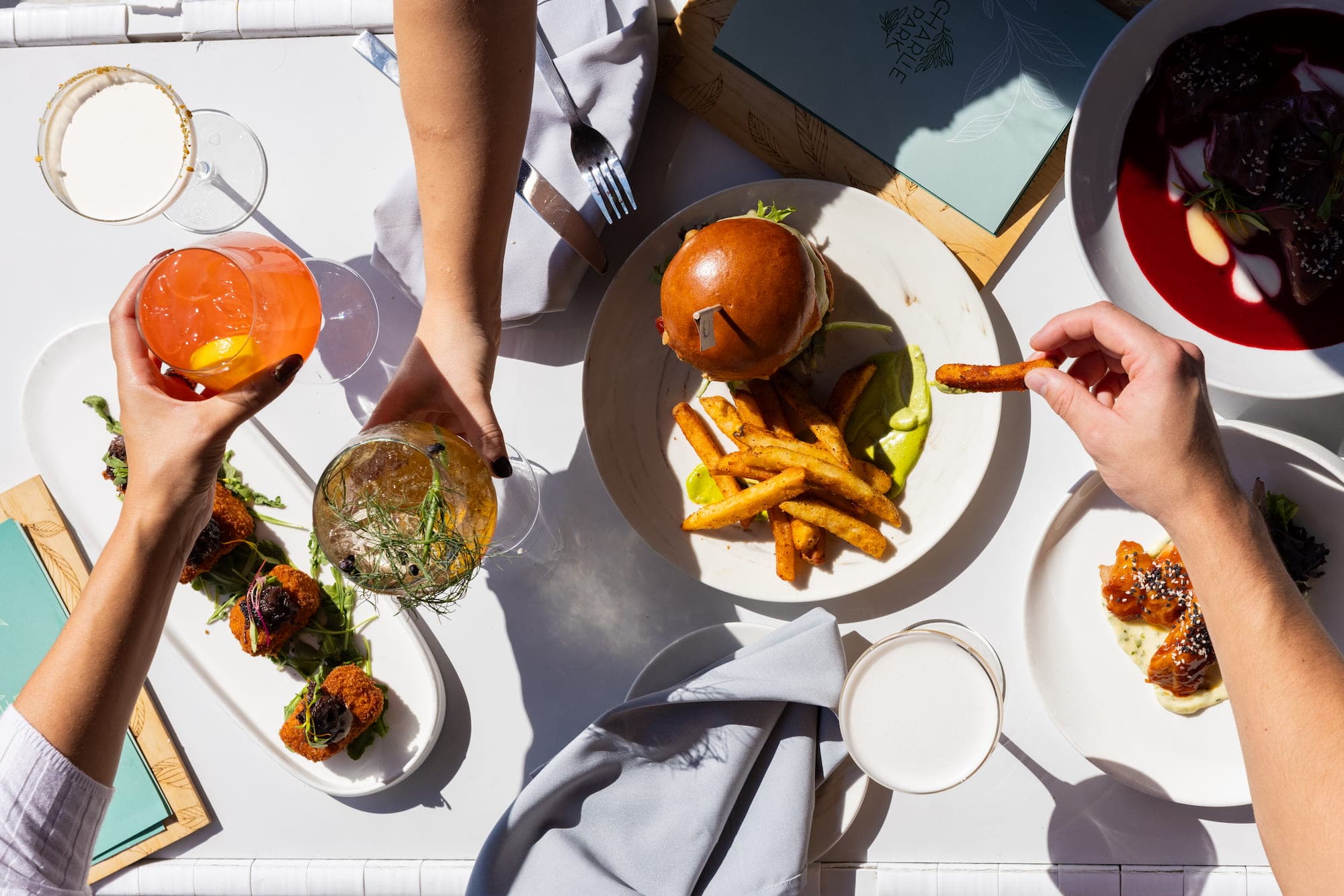 Hands reaching for cocktails, fries, crispy bites, and a beet dish spread across a table at Charlie Park