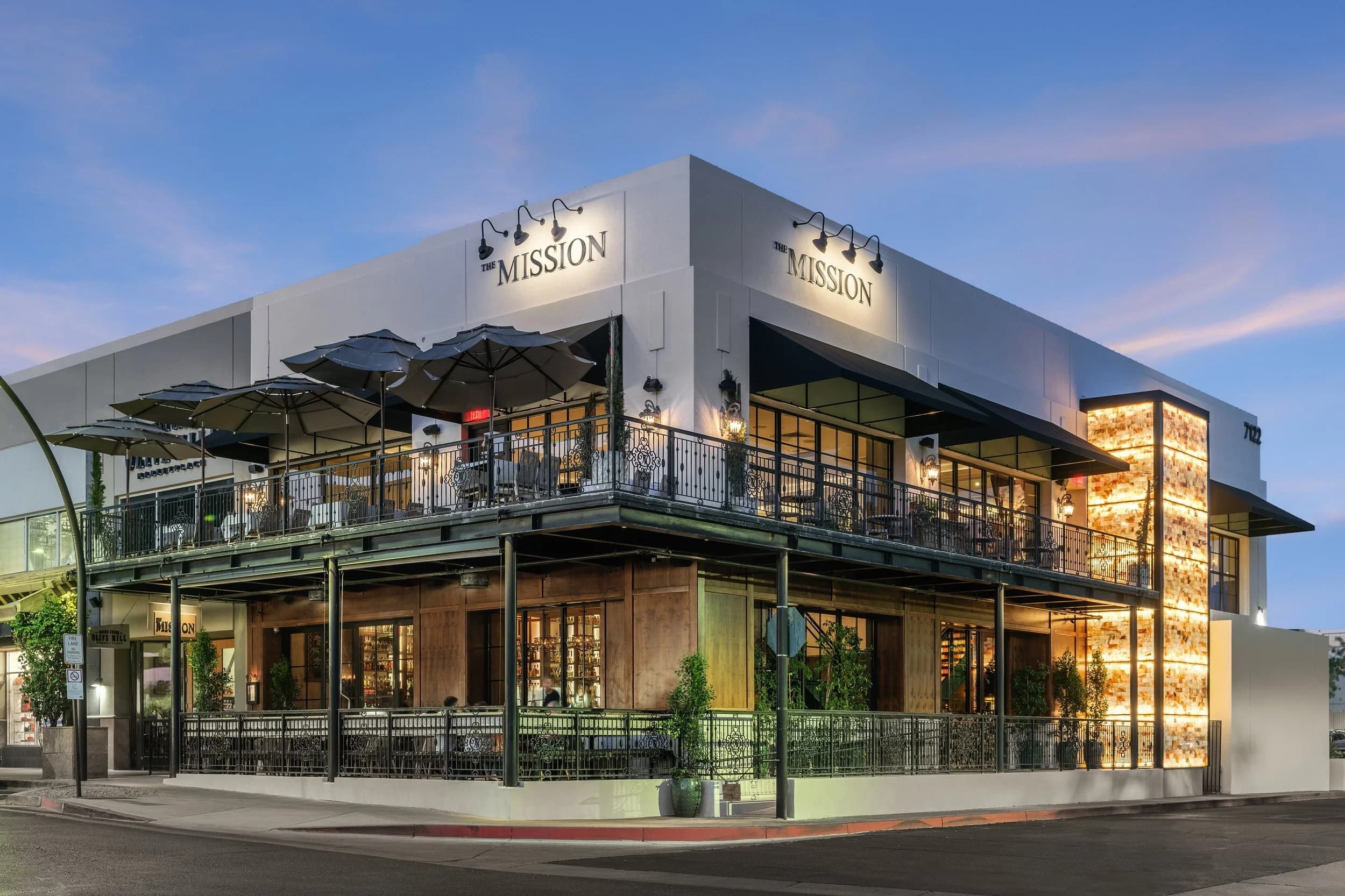 Twilight exterior of The Mission's two-story building with lit signage, patio dining, and open-air umbrellas