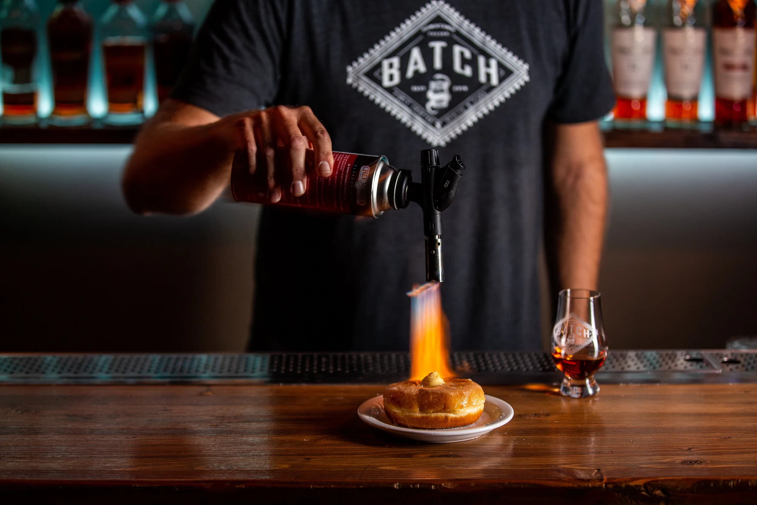A bartender in a Batch t-shirt torching the top of a flaming doughnut cocktail garnish over a whiskey glass at the bar