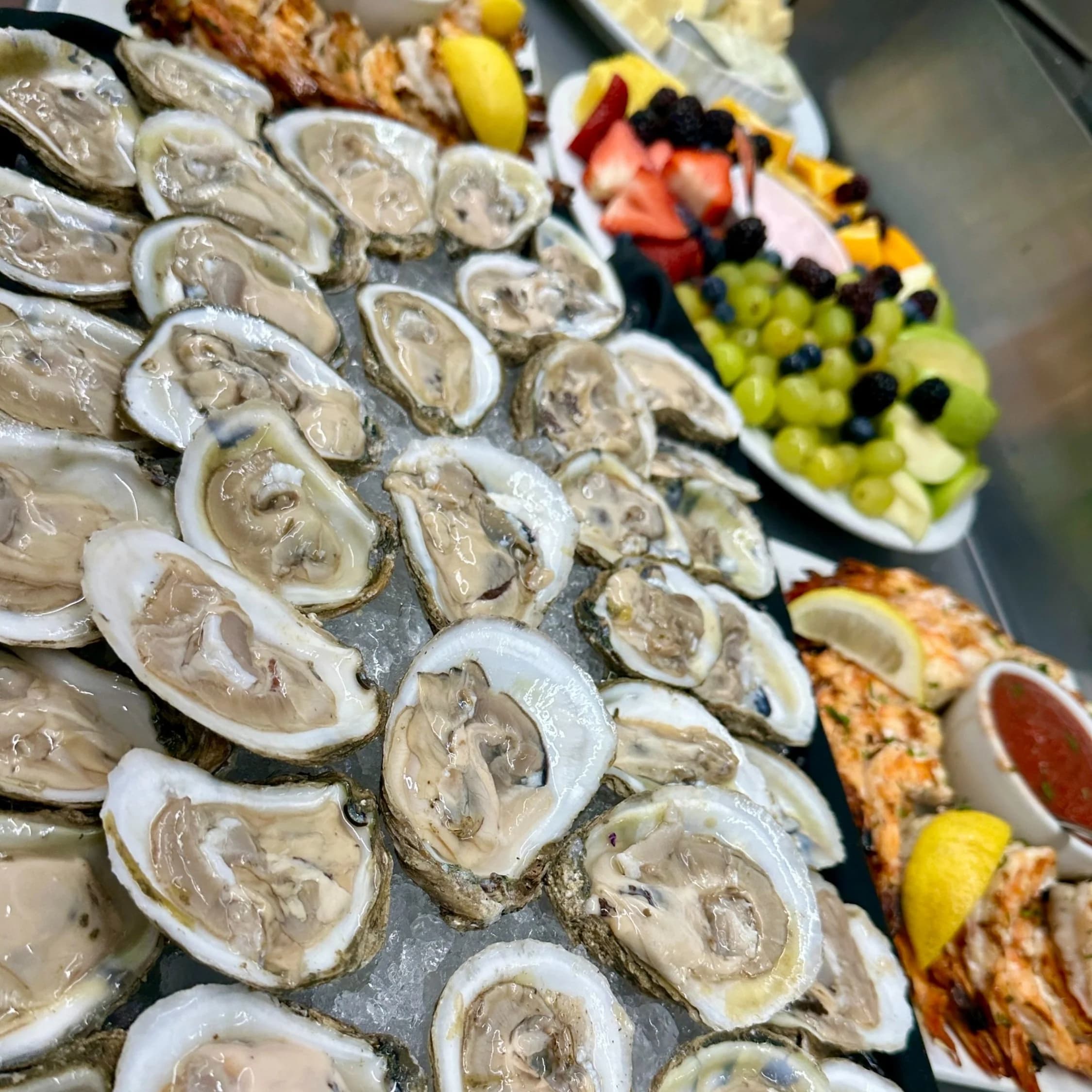 A large raw bar platter of freshly shucked oysters on ice with lemon wedges, alongside grilled shrimp and a fruit platter