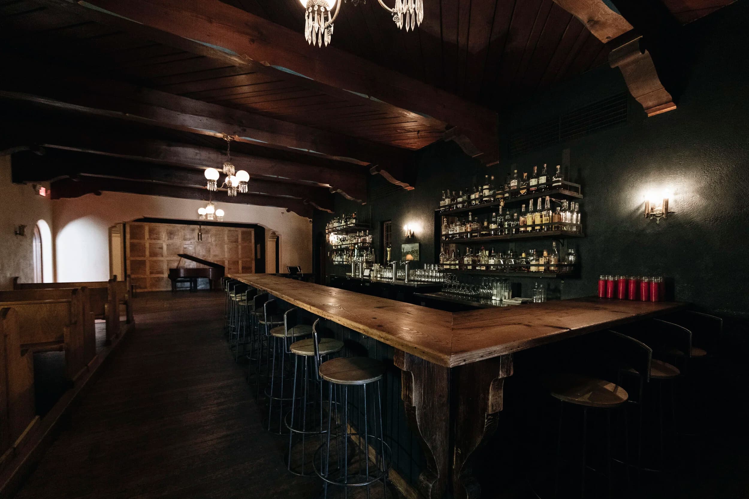 The Owls Club's Spanish-colonial bar room with candlelit tables, a piano, and pews along the wall under wooden beams