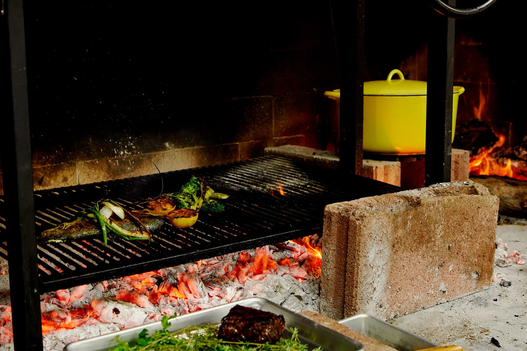 Tito & Pep's open mesquite fire pit and grill, vegetables and a steak searing over the coals, a yellow pot warming on the fire